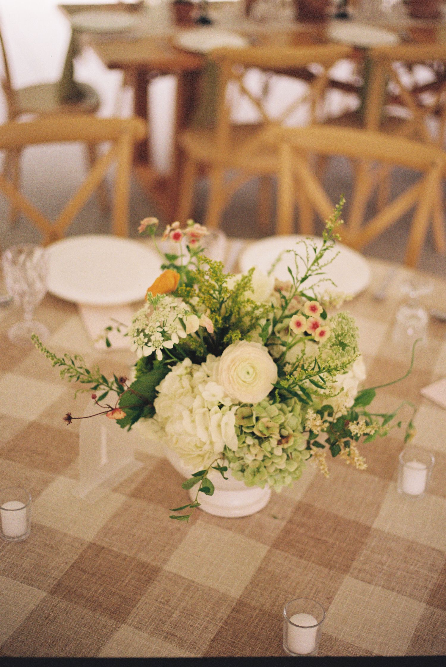 White hydrangea and ranunculus floral centerpiece in white vase on plaid tablecloth with candles.
