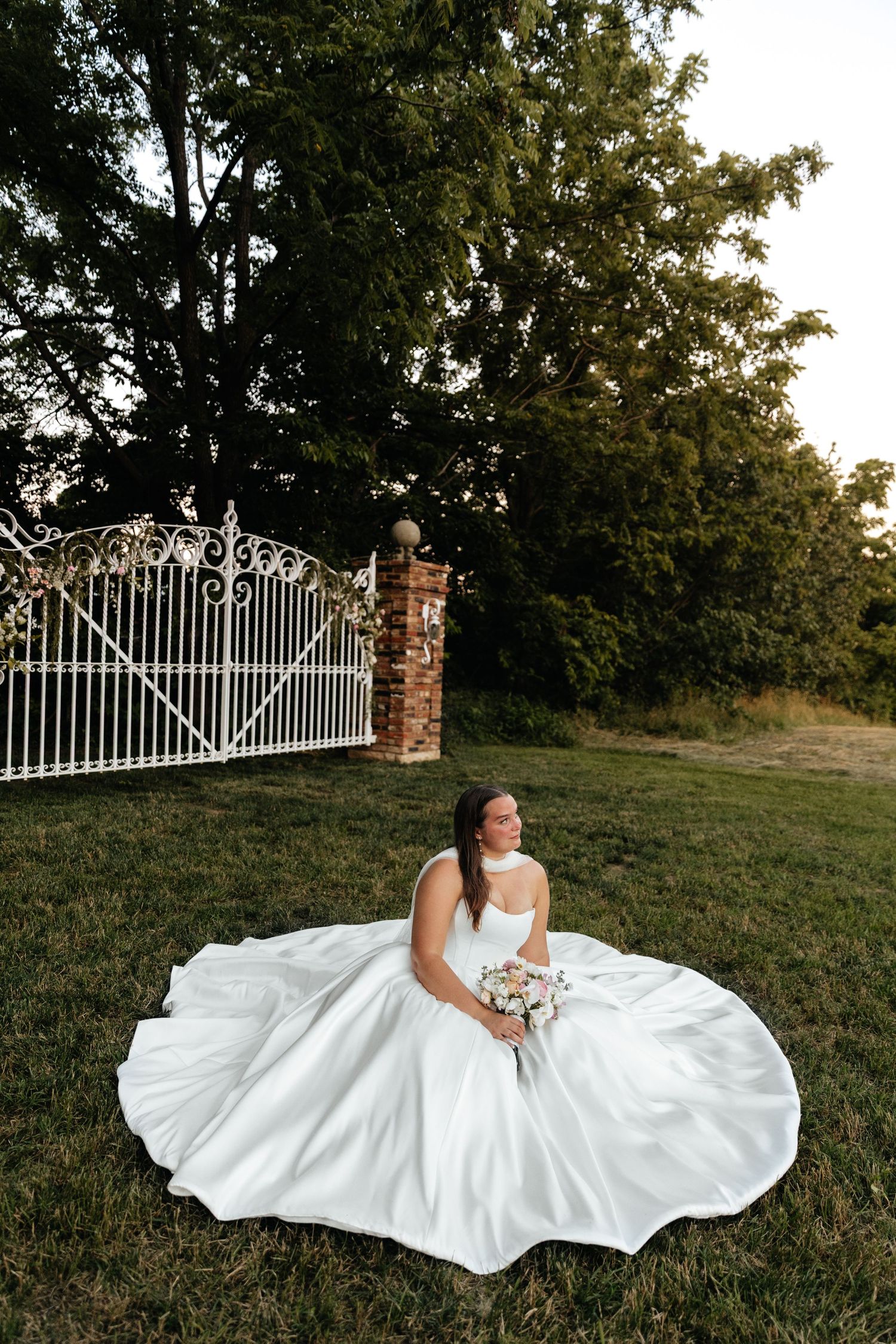 Stunning white wedding dress spreads in full circle on grassy hill by decorative gates.