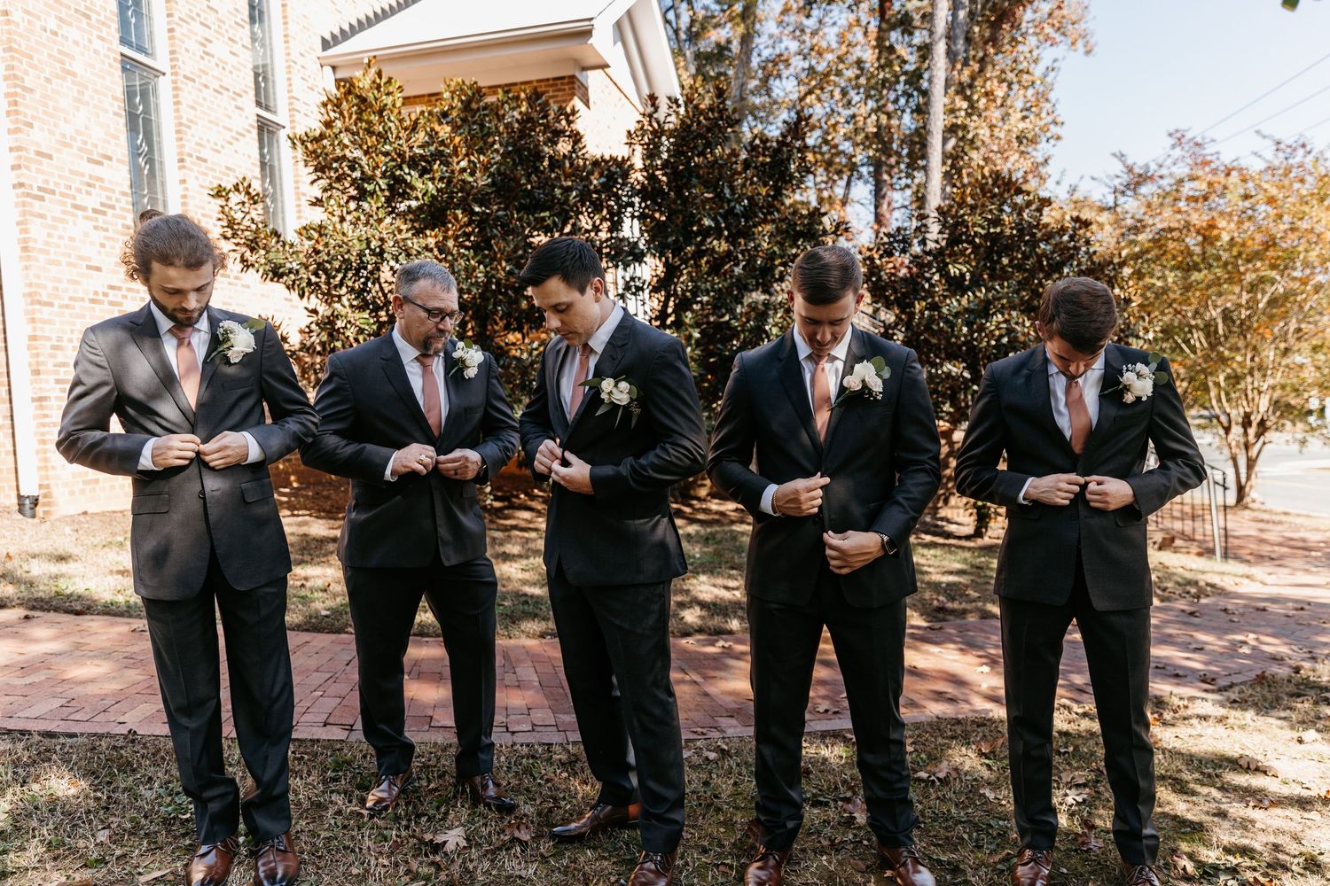 Groomsmen in black suits checking their phones while standing outside.