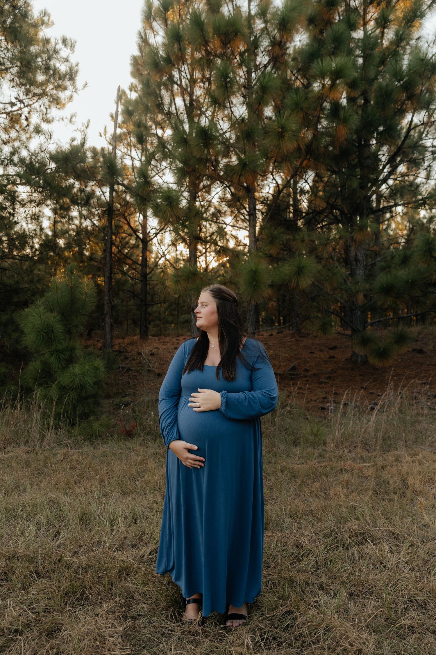 Expectant mother in blue dress standing in wooded area during sunset.