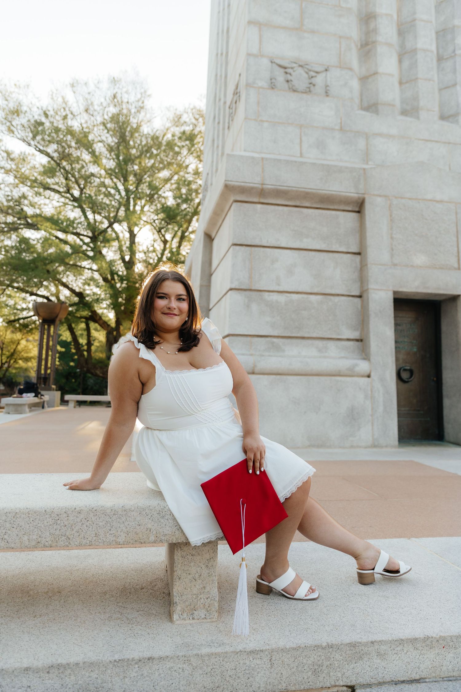 Graduate in white dress posing on stone steps with red graduation cap.