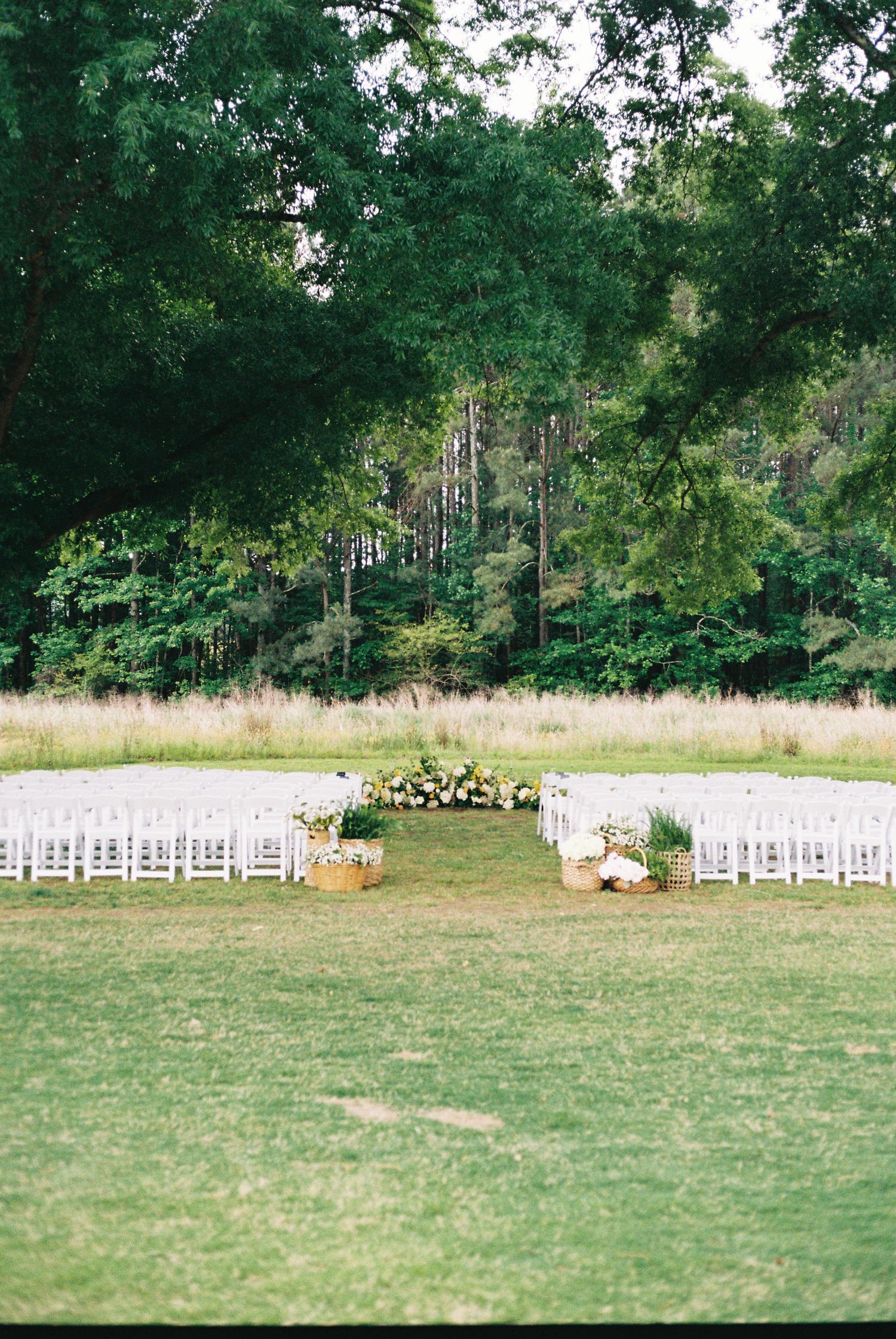 Outdoor wedding ceremony setup with white chairs, floral altar arrangements, and lush green trees in a natural meadow setting.