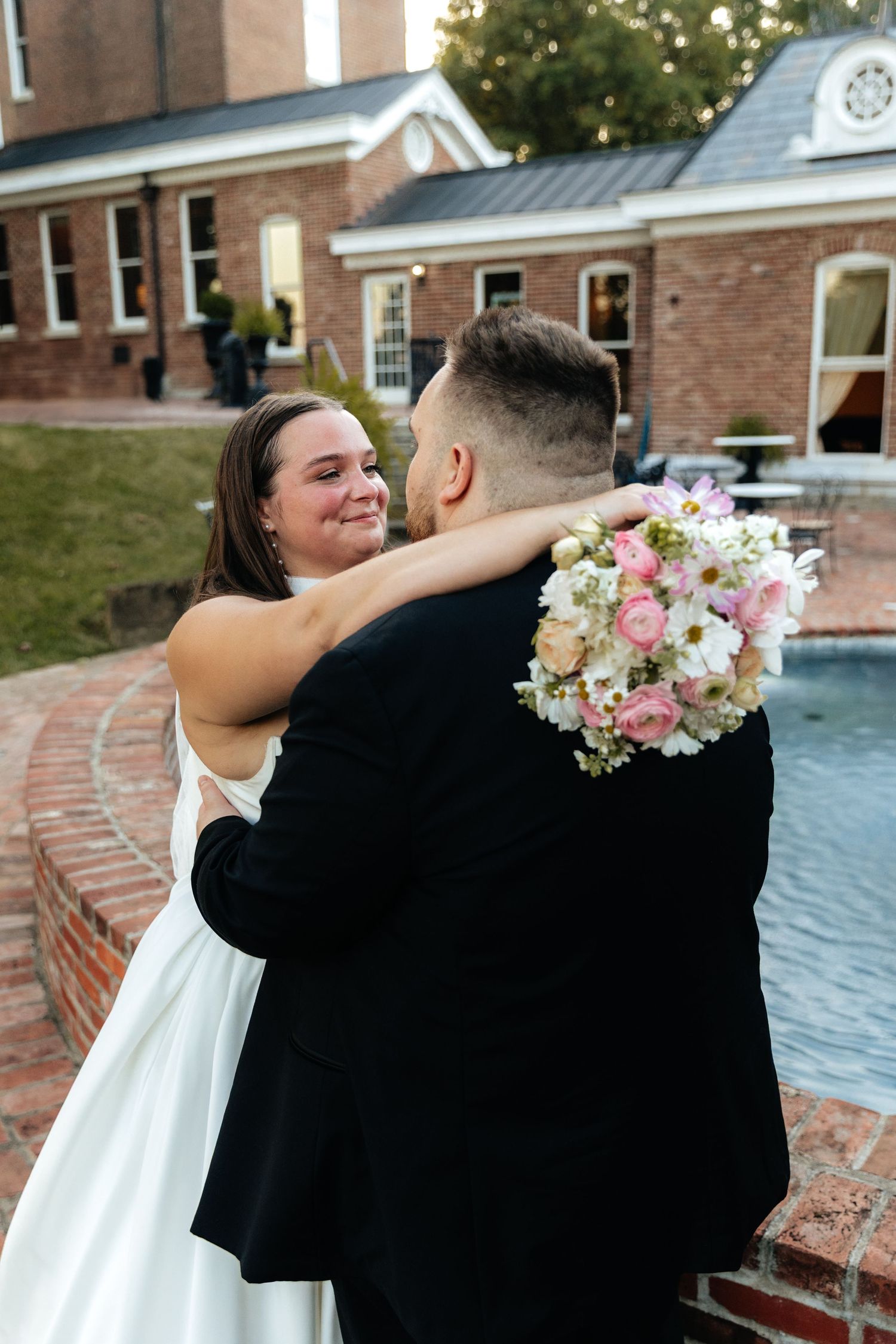 Couple embraces near a brick building with pink floral bouquet.