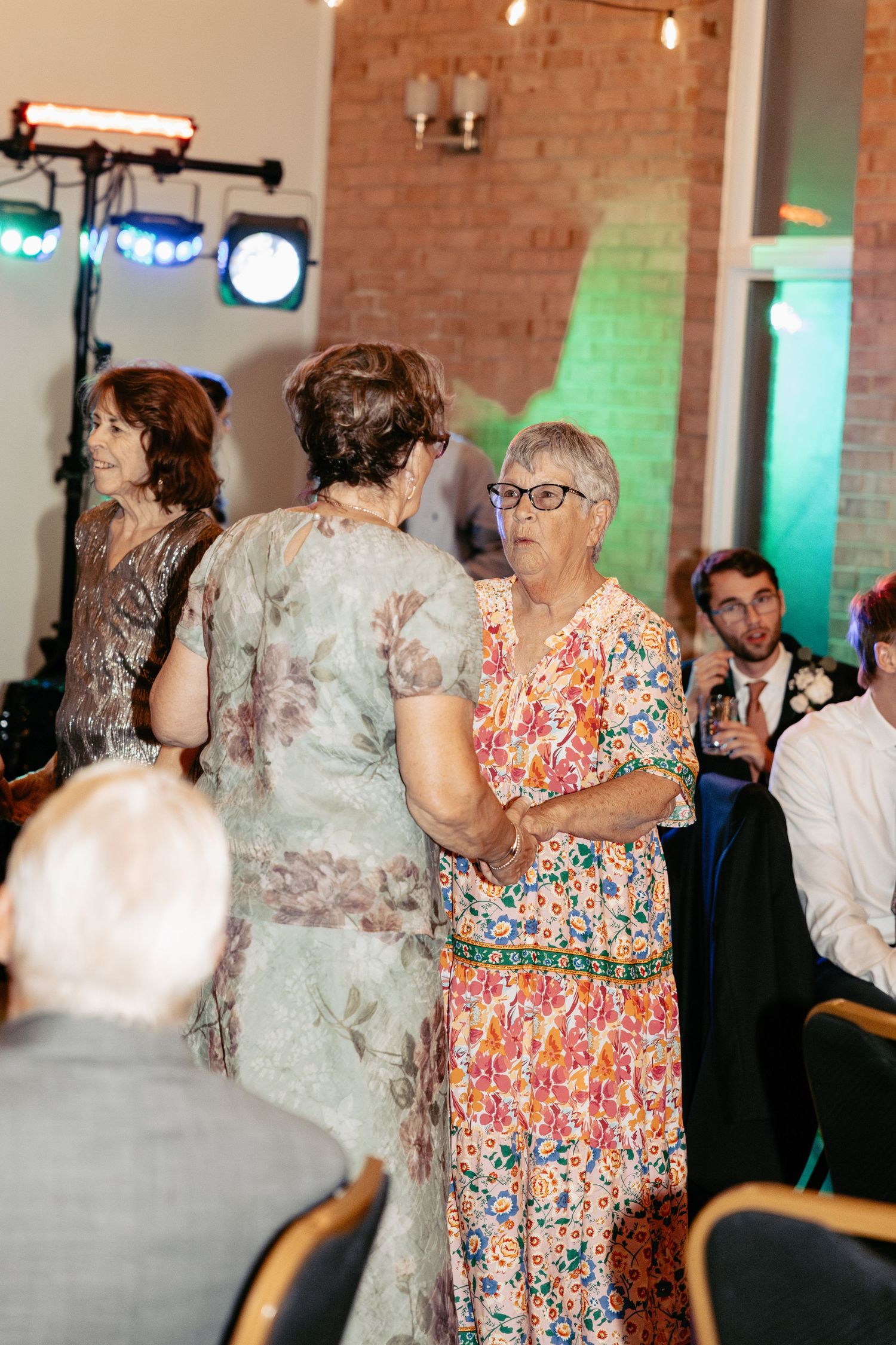 Wedding guests enjoying conversation during reception in venue with exposed brick walls and colorful lighting.