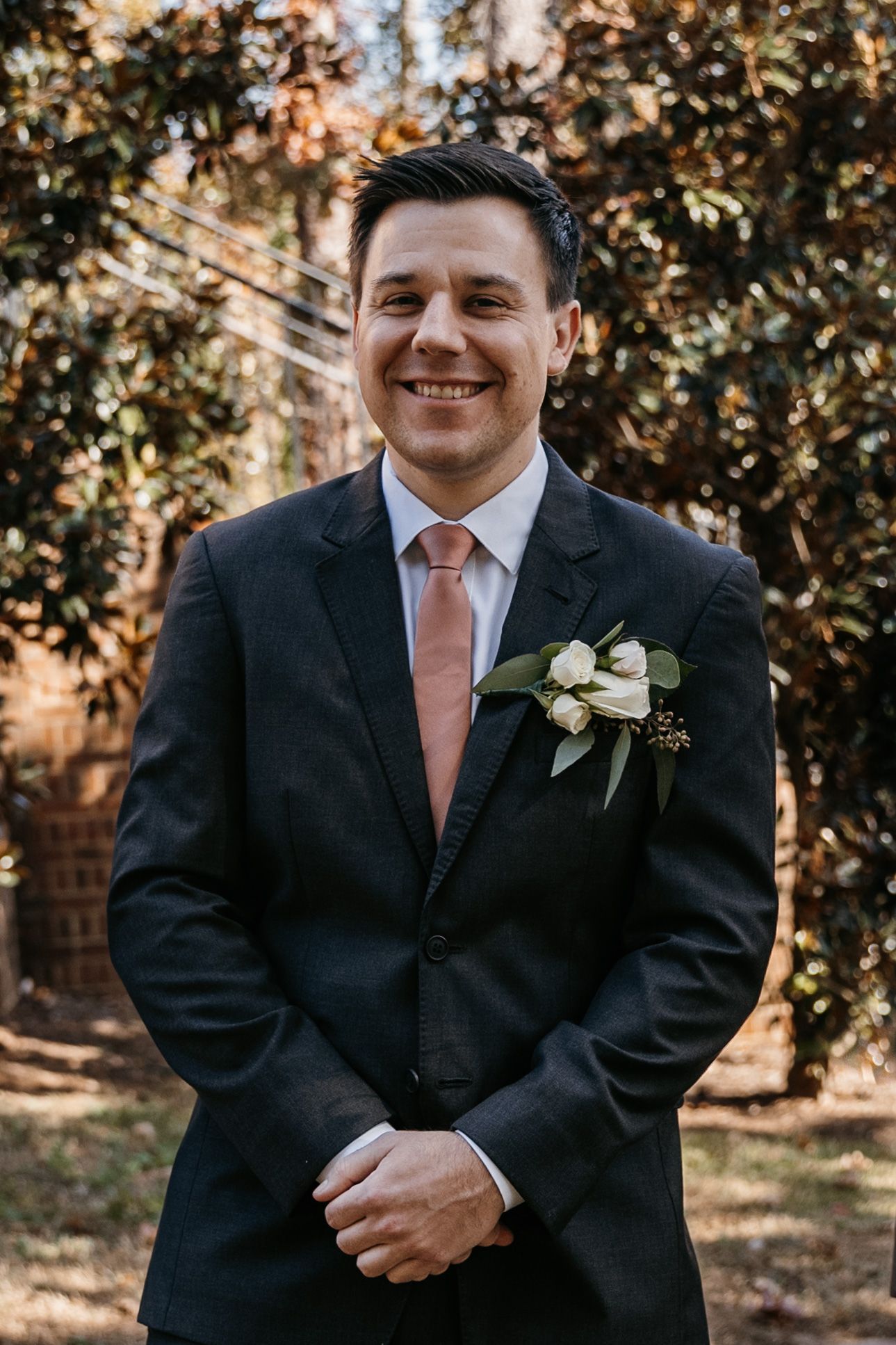 Wedding portraits of groom in black suit with coral tie against natural outdoor backdrop.