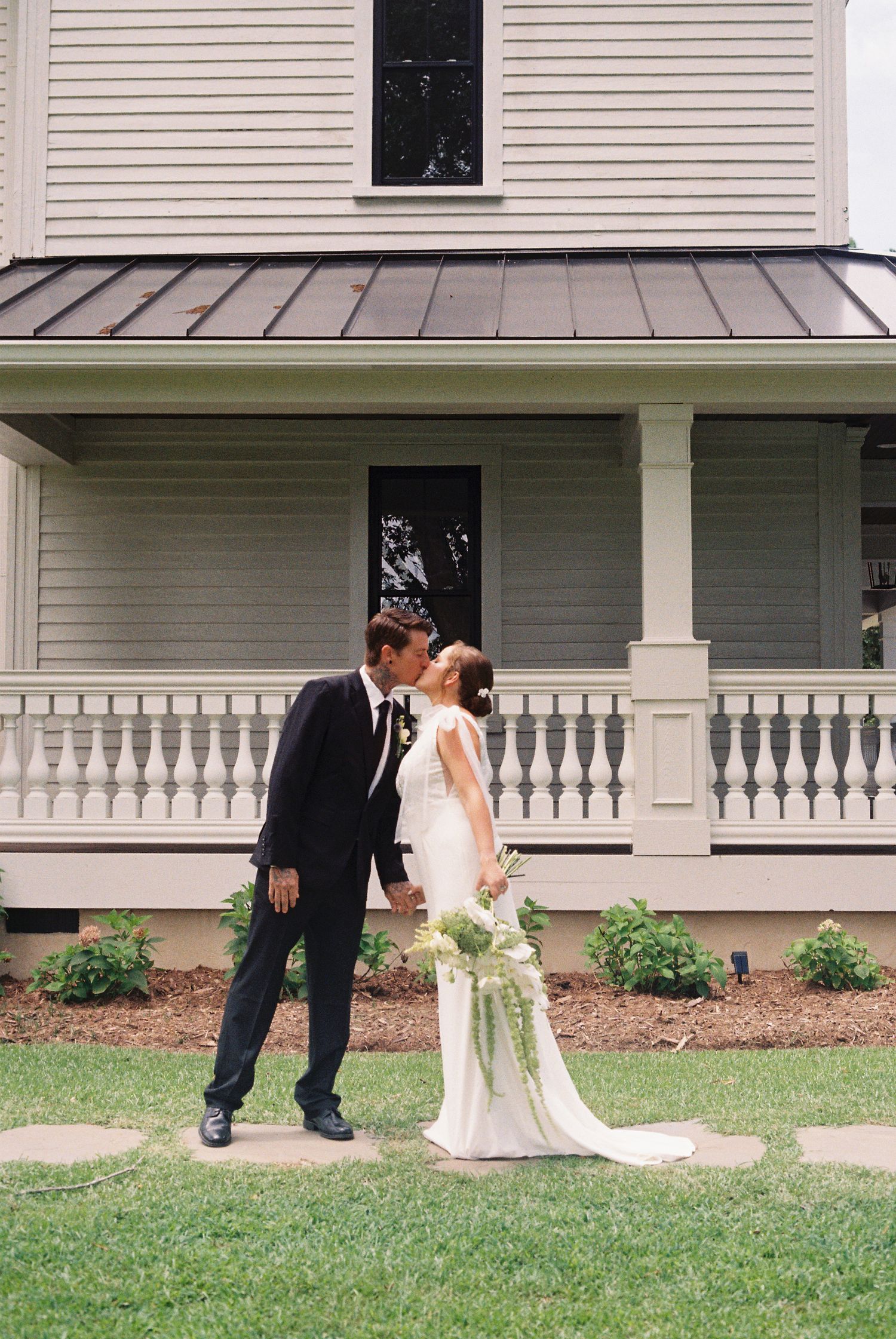 Wedding couple posing on the lawn in front of a white colonial-style house.