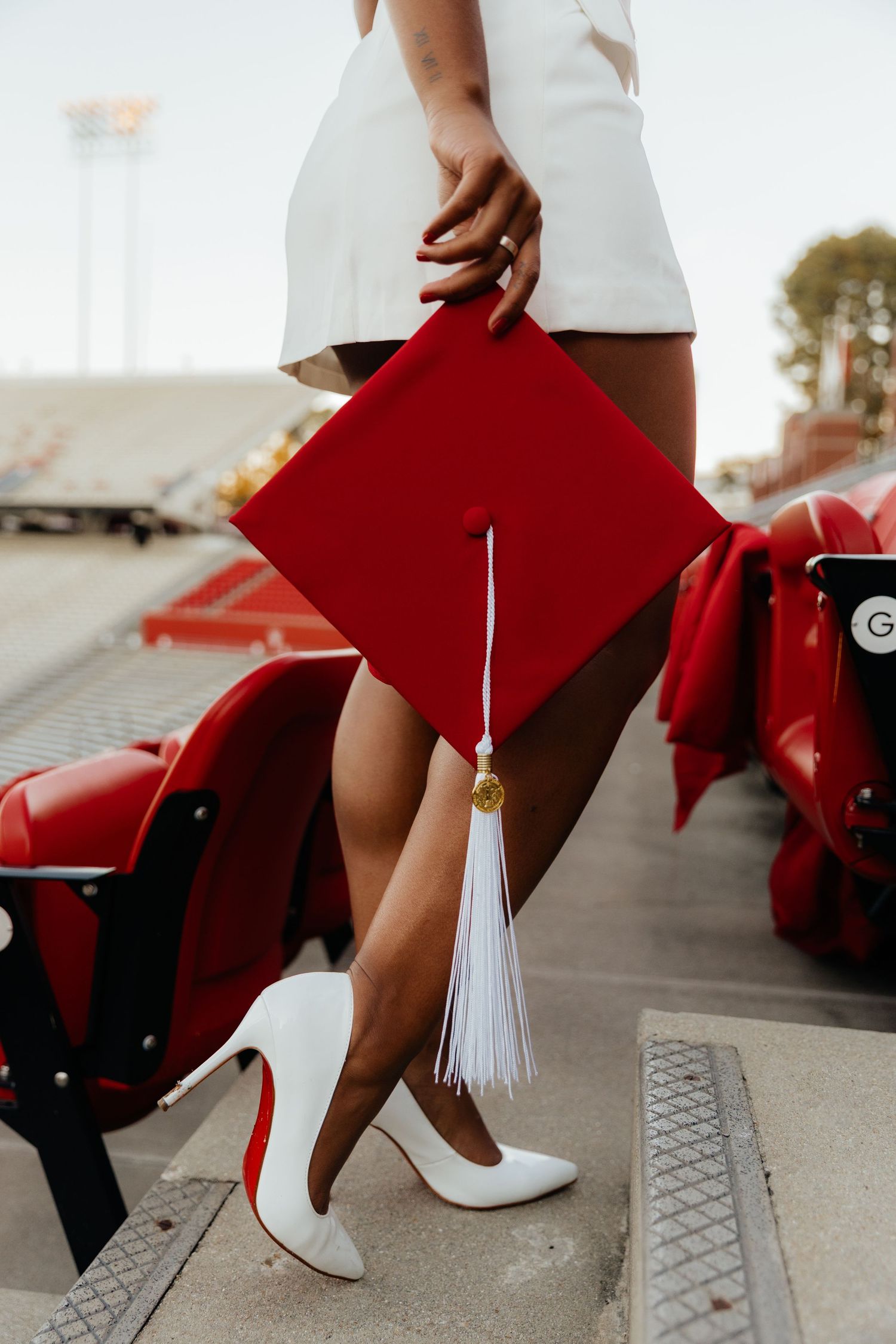 Close-up of graduate holding red mortarboard cap wearing white heels at stadium graduation photos.