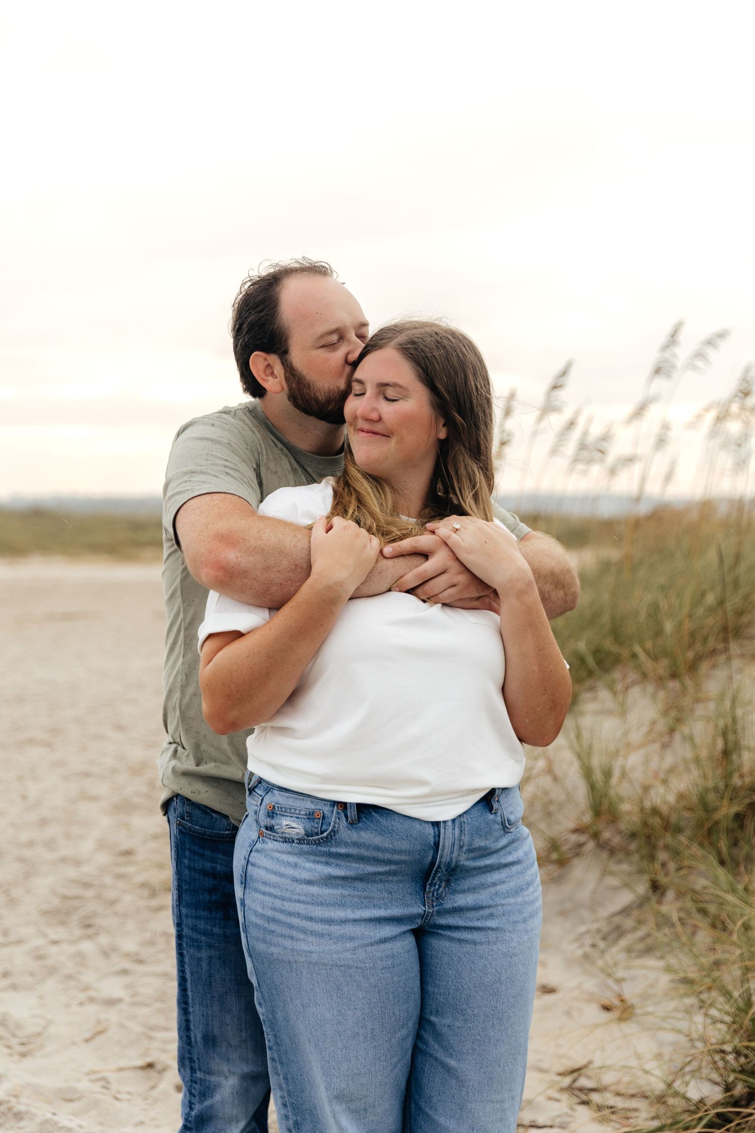 Romantic couple embraces on a sandy beach with tall grass in the background during golden hour.