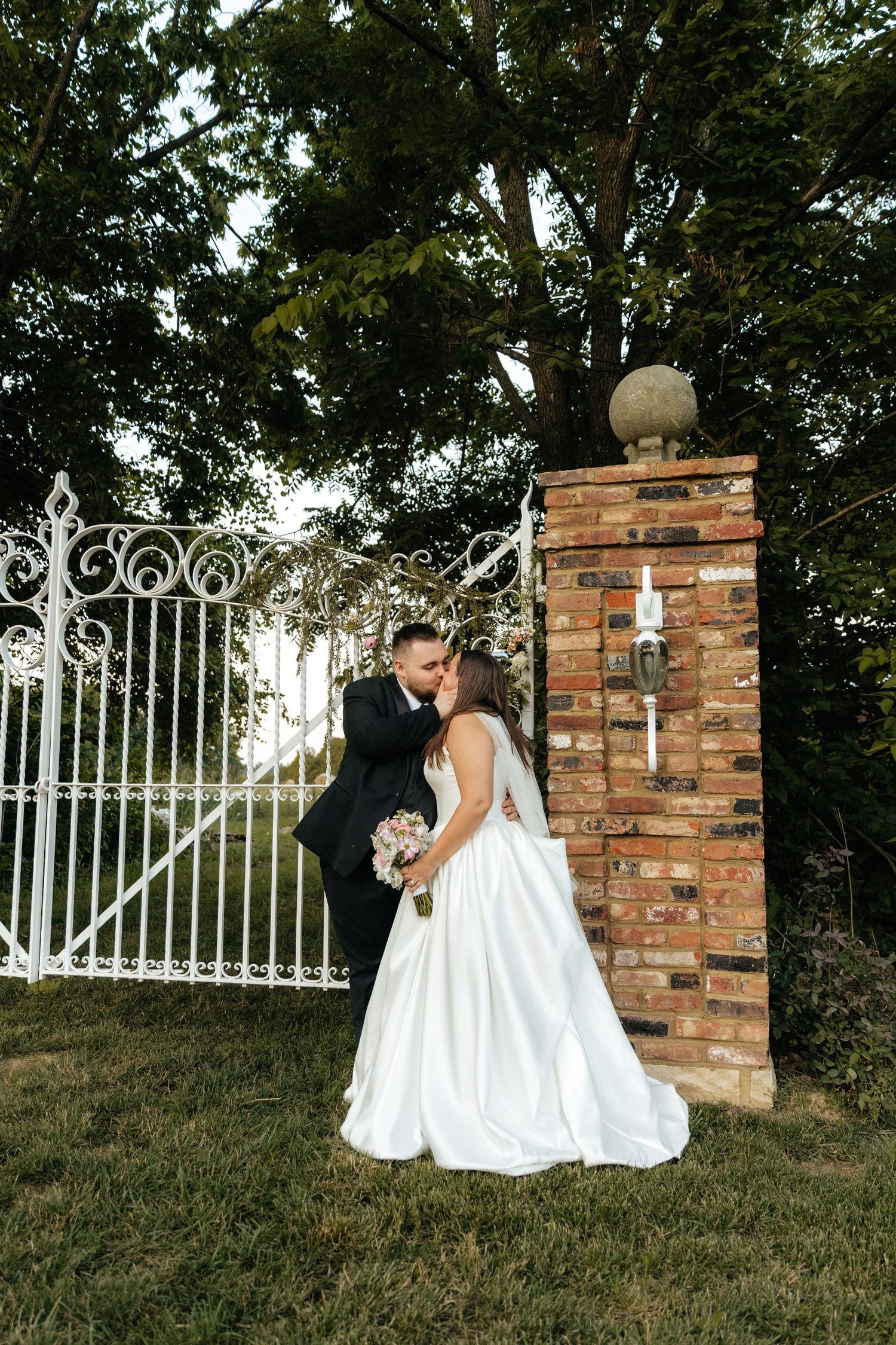 Couple poses near white iron gate and brick pillars in garden setting.