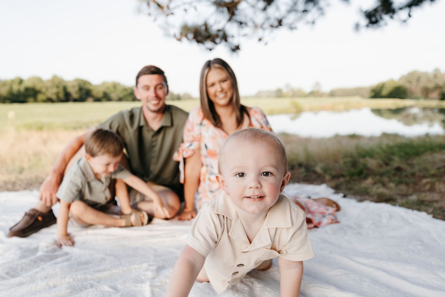 Family relaxing together on a white blanket by a pond on a sunny day.