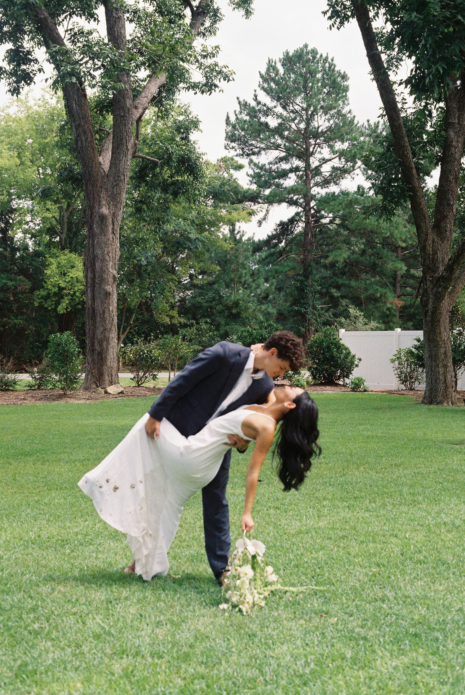 Romantic wedding couple poses in a series of dips on a grassy lawn with trees in the background.