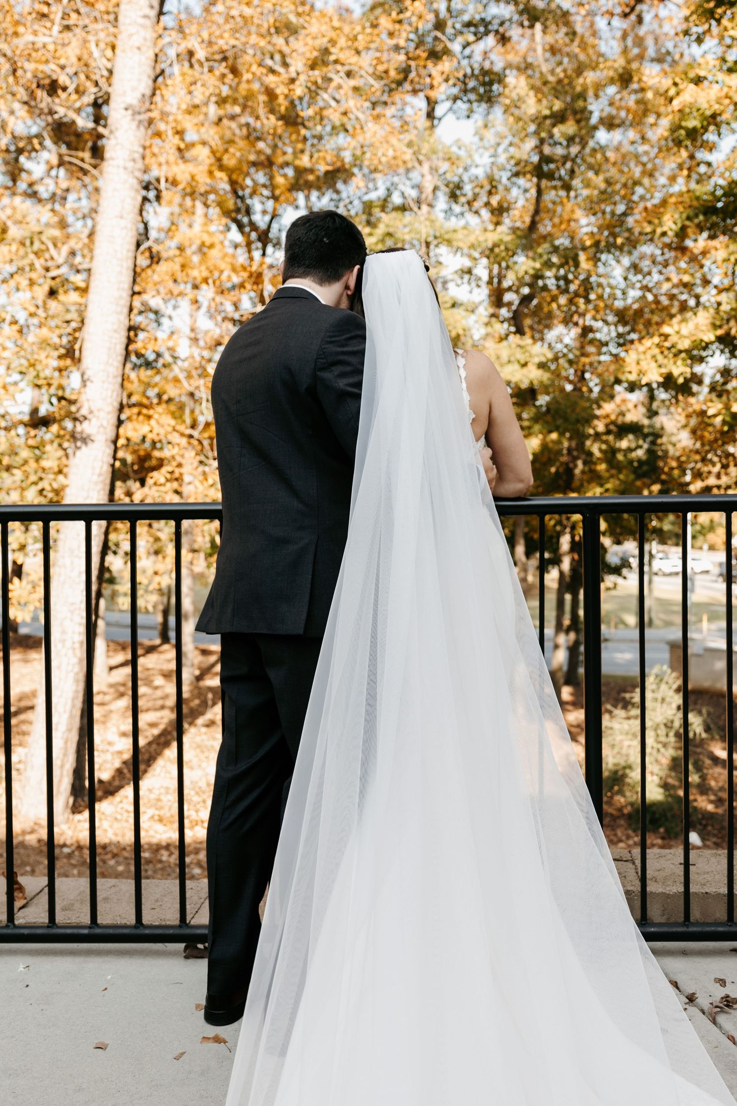 Wedding couple photographed from behind against autumn trees and metal railing.