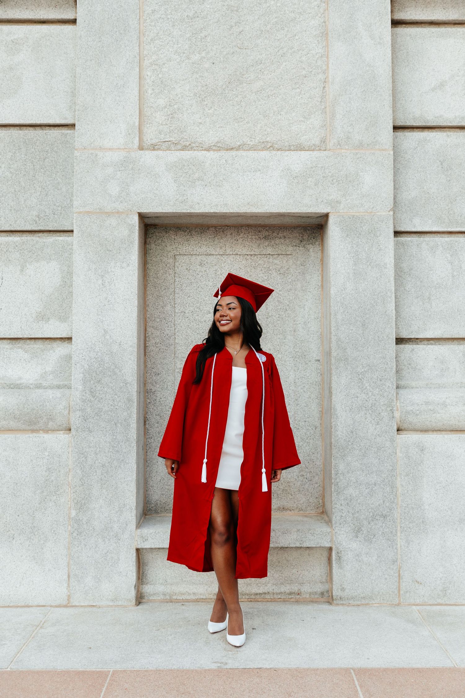 Graduate in red cap and gown standing in stone building alcove wearing white dress.