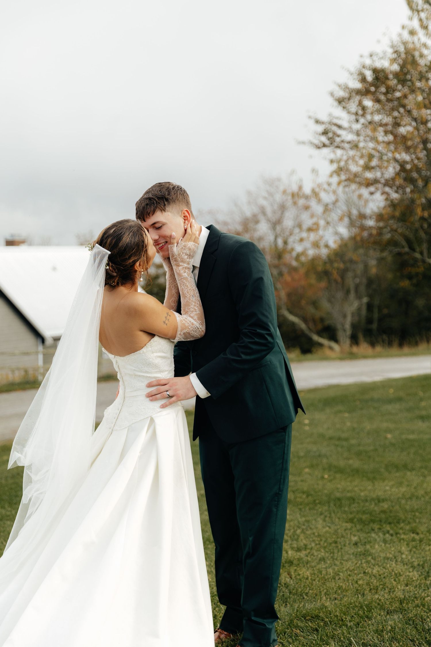 A bride and groom share an intimate moment outdoors, she in a white gown with veil, he in a dark suit.
