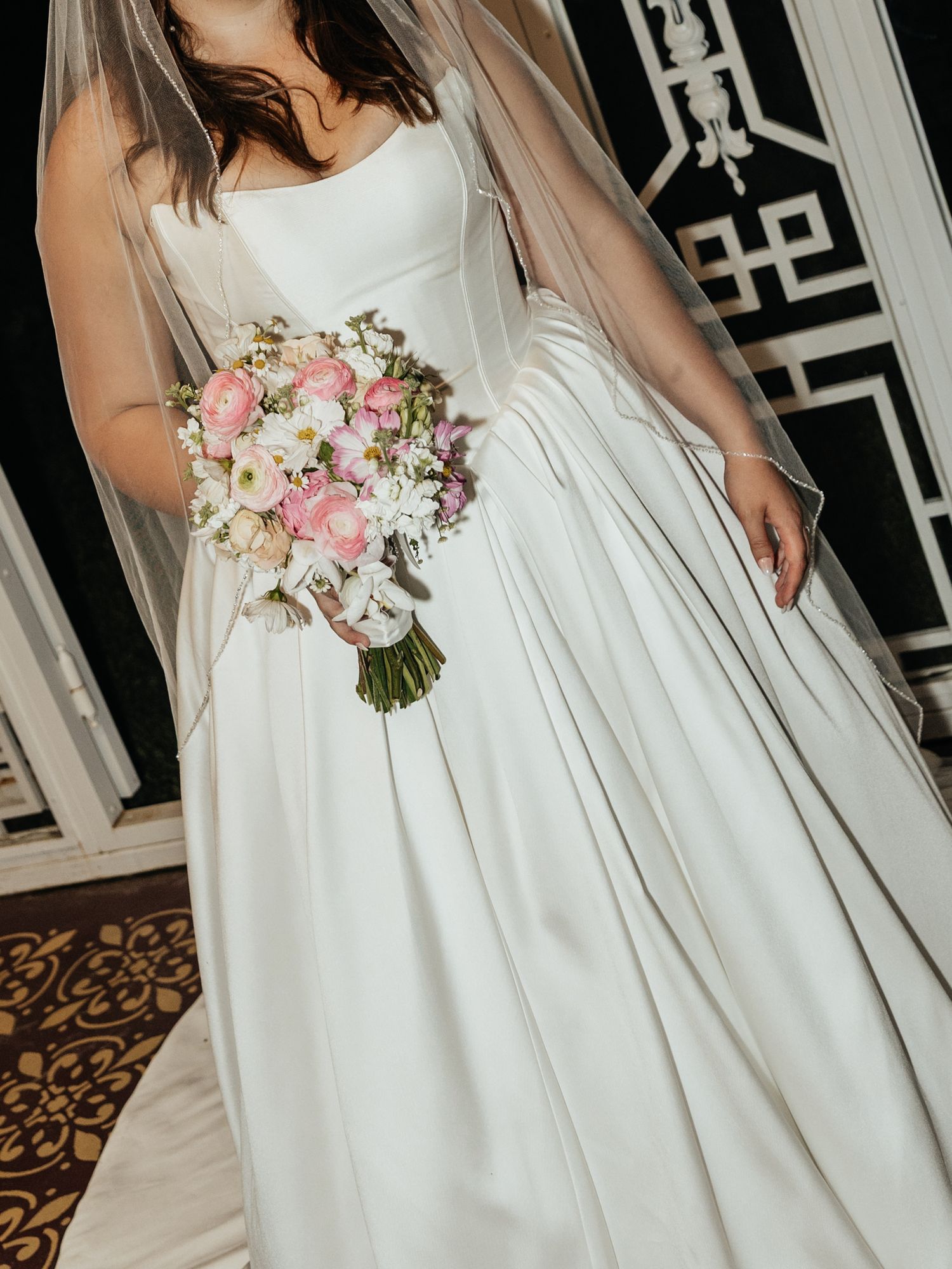Close-up of white wedding dress details with pink floral bouquet against ornate background.