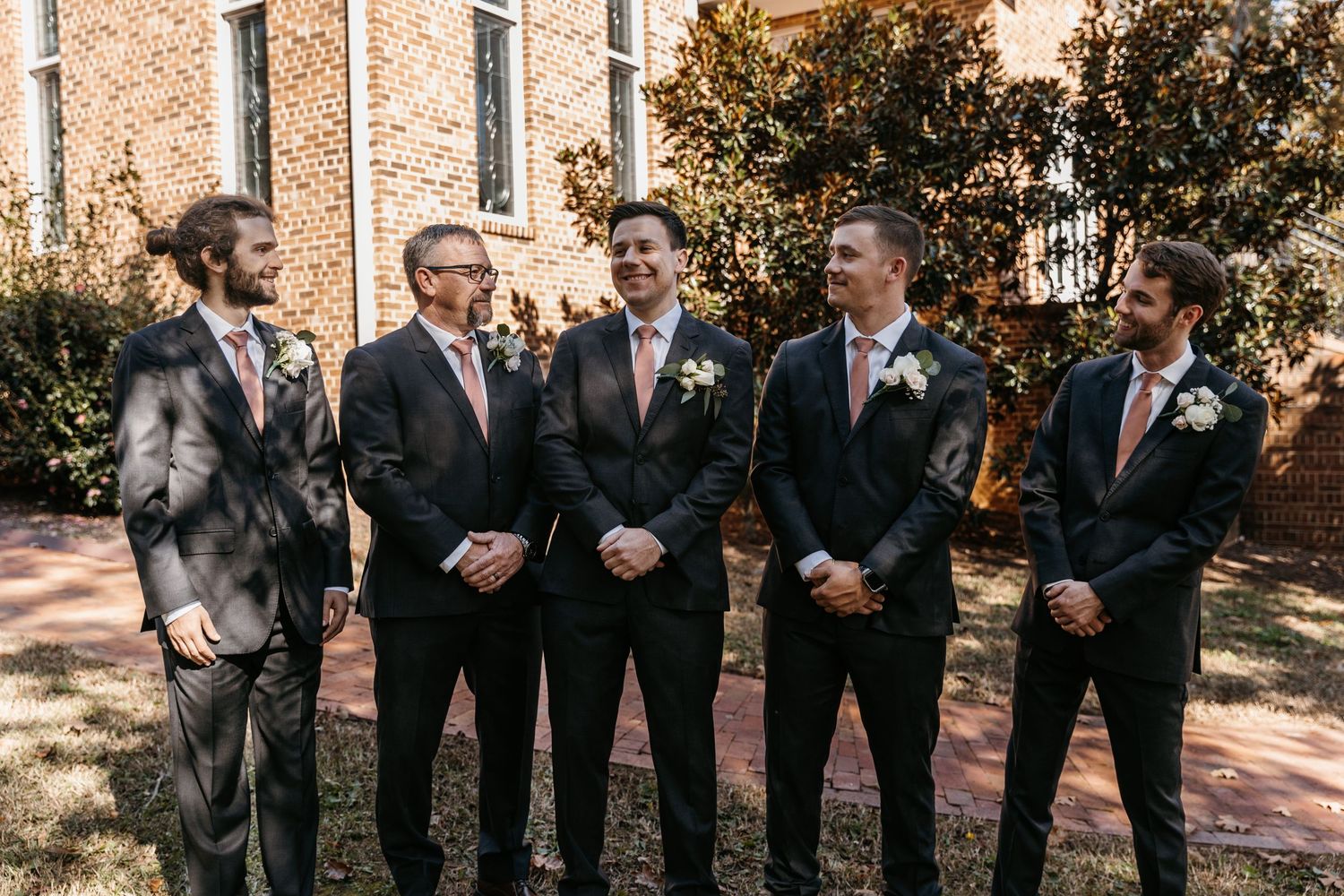 Groomsmen in dark suits with coral ties standing together outdoors.