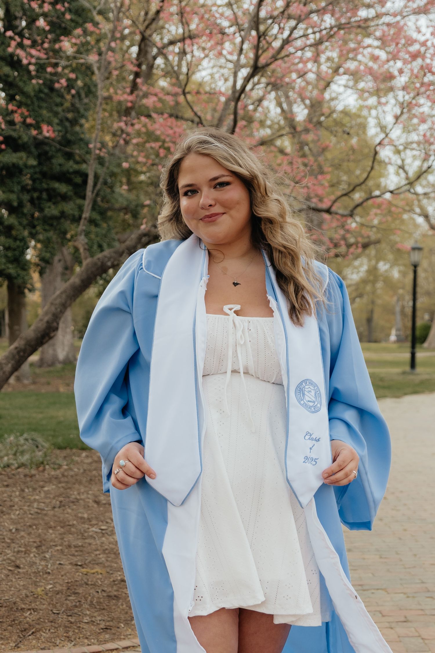 Young woman in light blue graduation gown and stole smiling outdoors near blooming trees on campus.