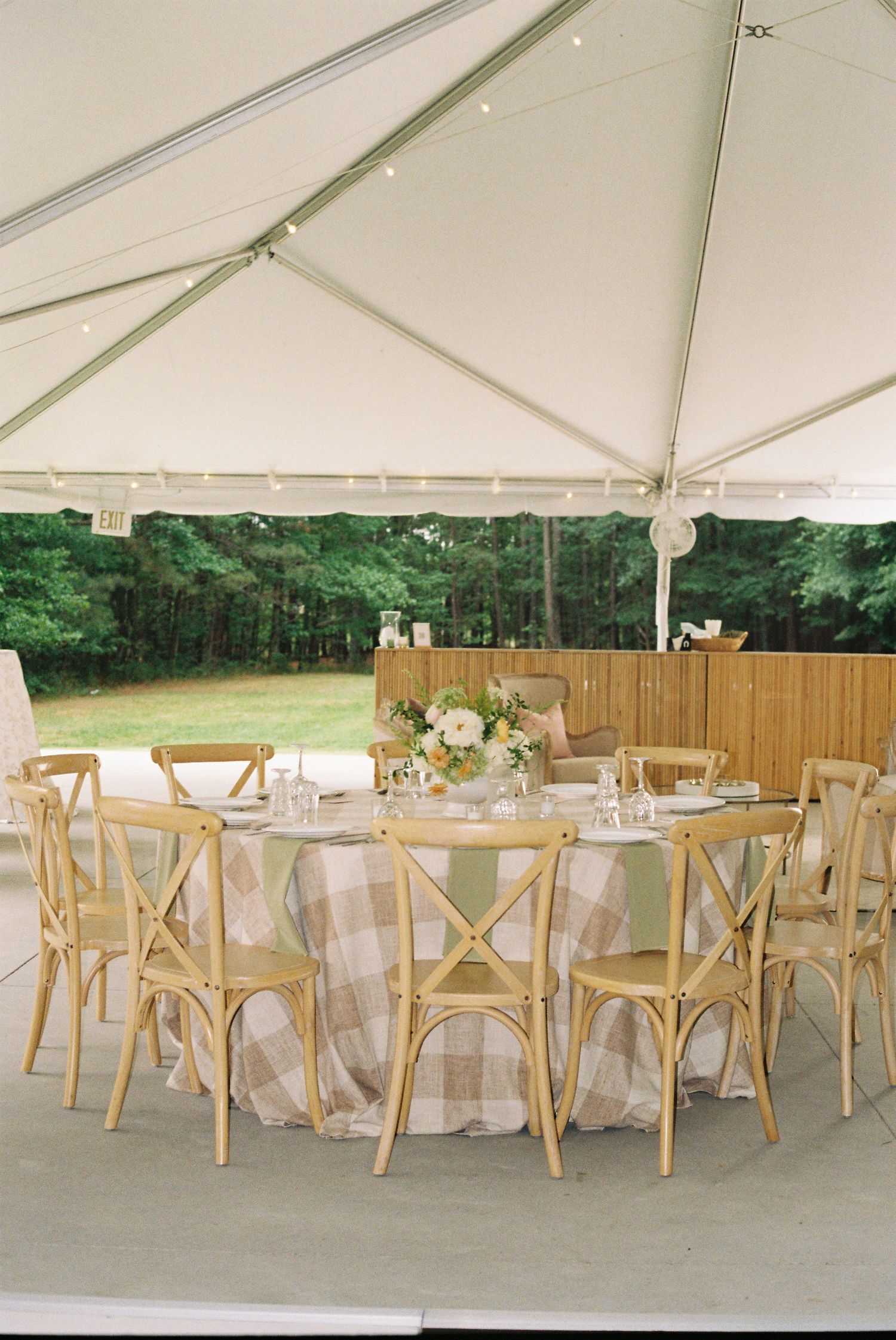 Elegant outdoor wedding reception under a white tent with wooden cross-back chairs and plaid tablecloth centerpiece.