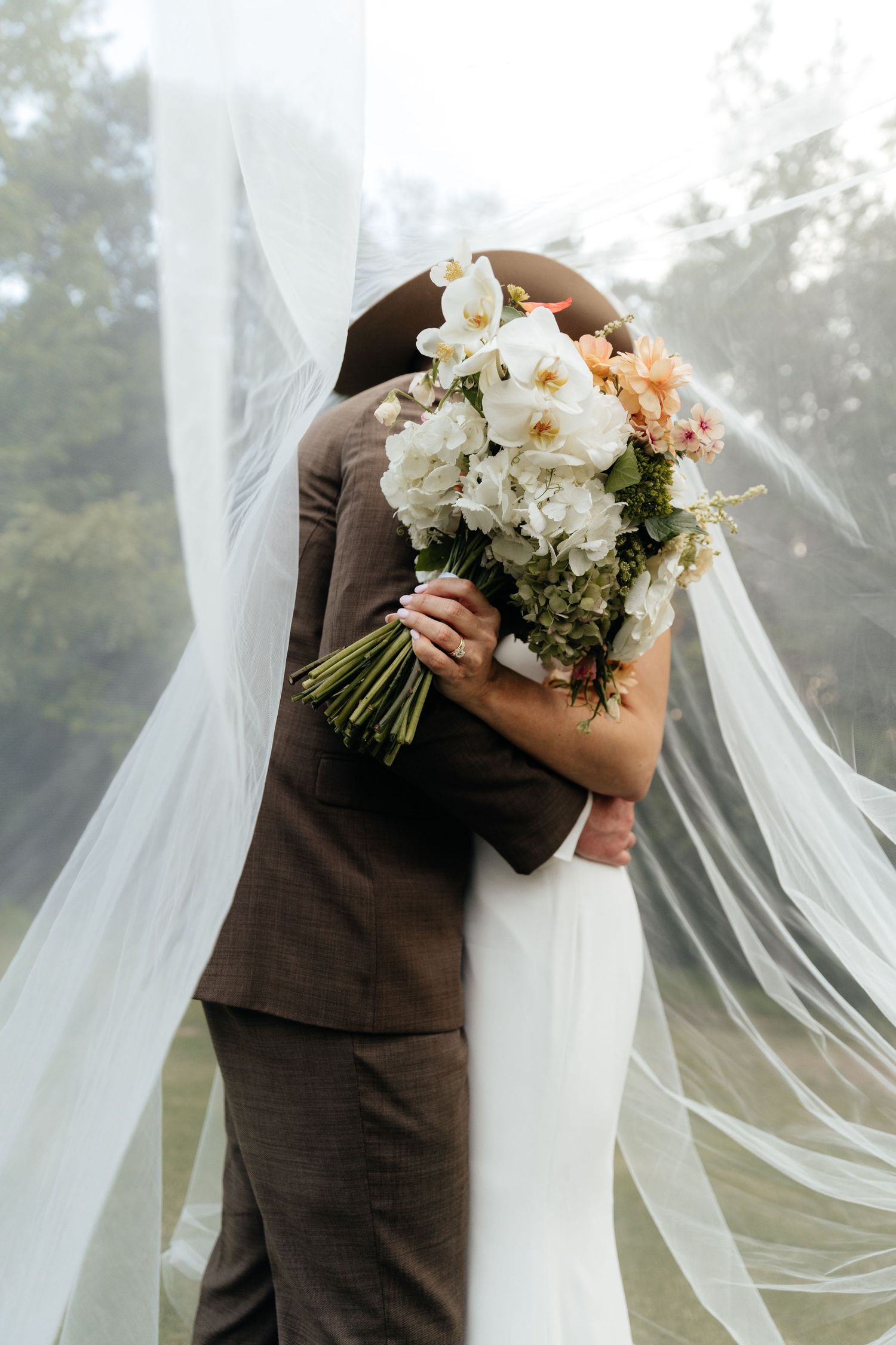 Bride and groom embracing behind a white floral bouquet, with a flowing veil and soft natural light.