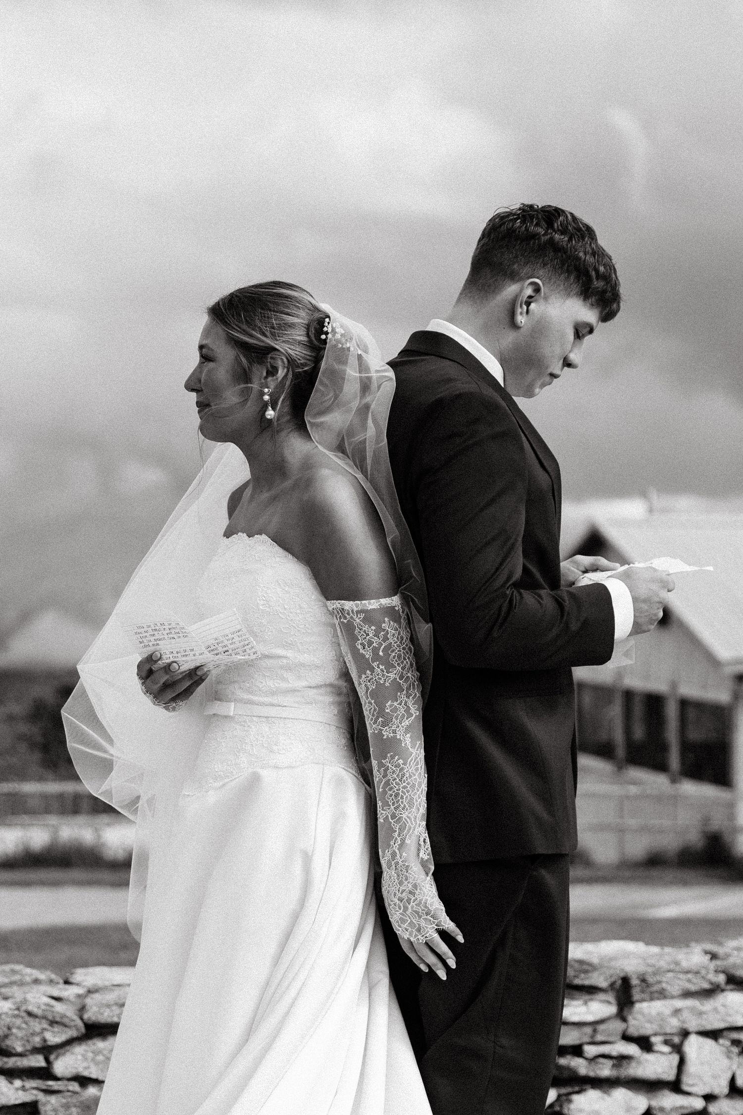 Black and white photo of bride and groom standing back-to-back reading vows outdoors on their wedding day.