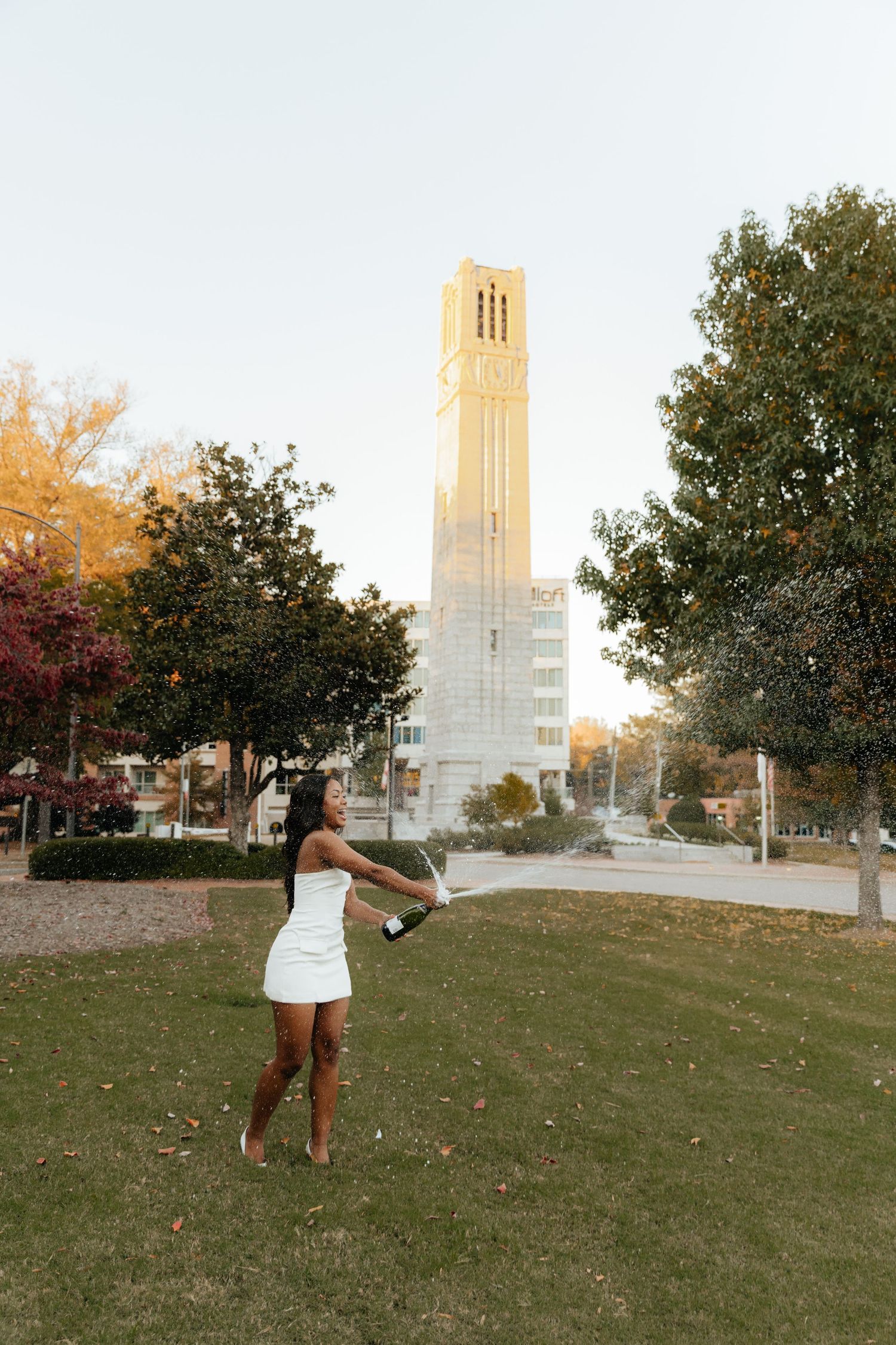 A graduate in white dress stands on grass with a bell tower in the background during sunset.