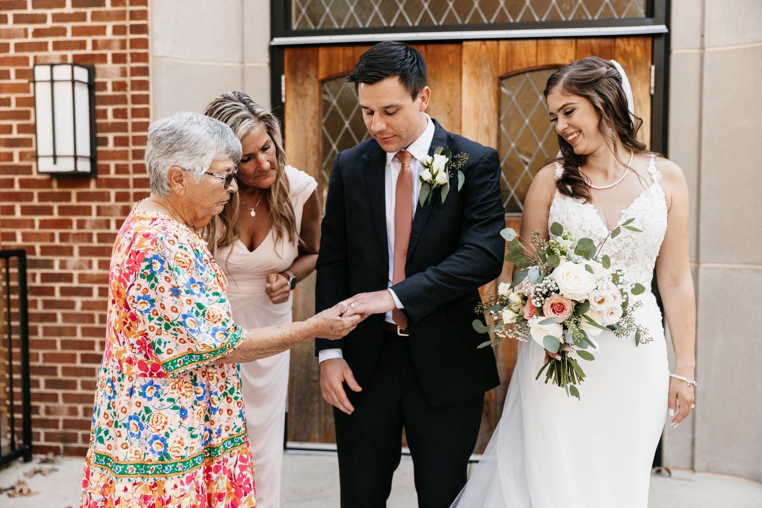 Wedding party gathers outside a traditional brick church entrance for formal photos.