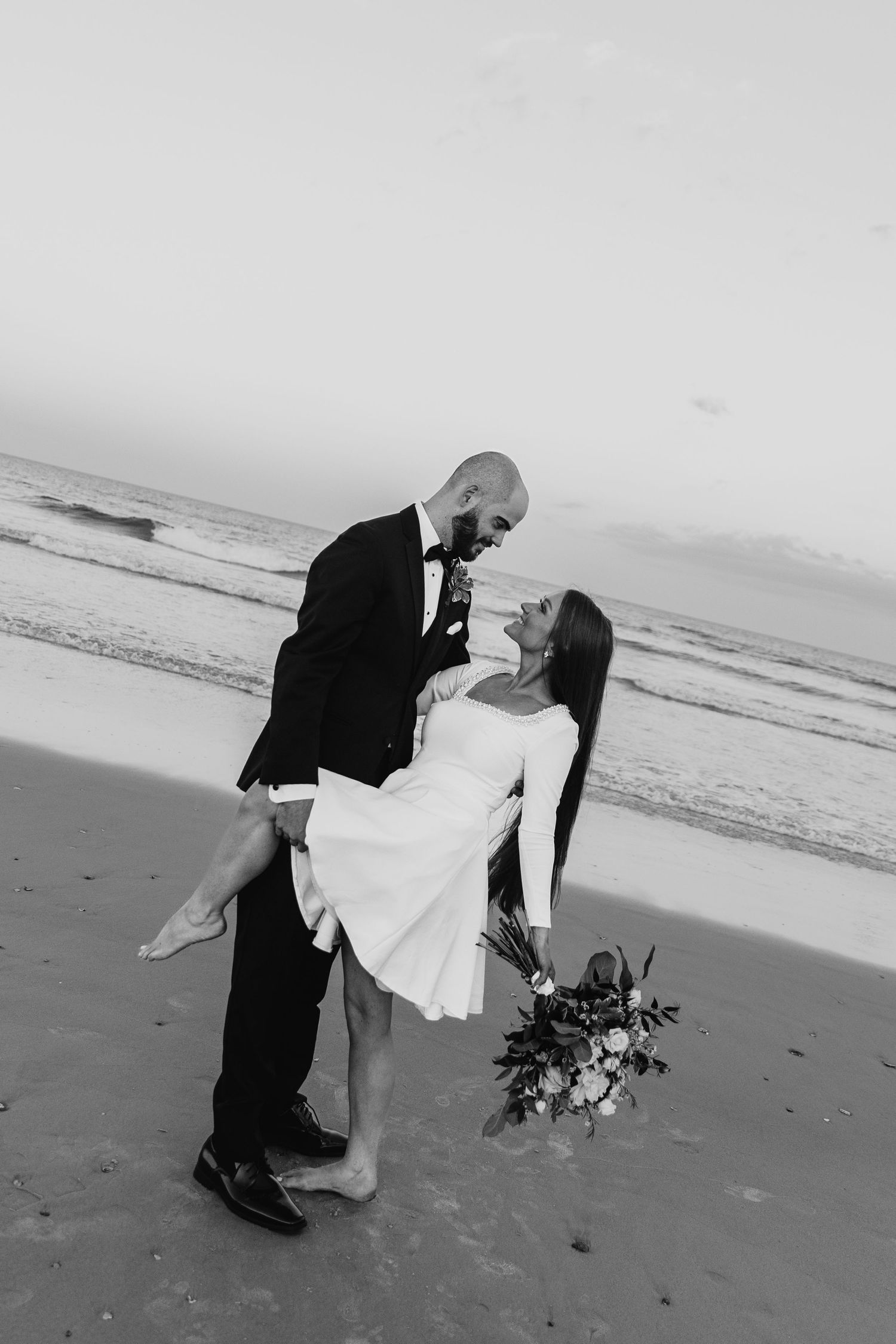 A black and white beach wedding photo capturing a romantic dip kiss moment.