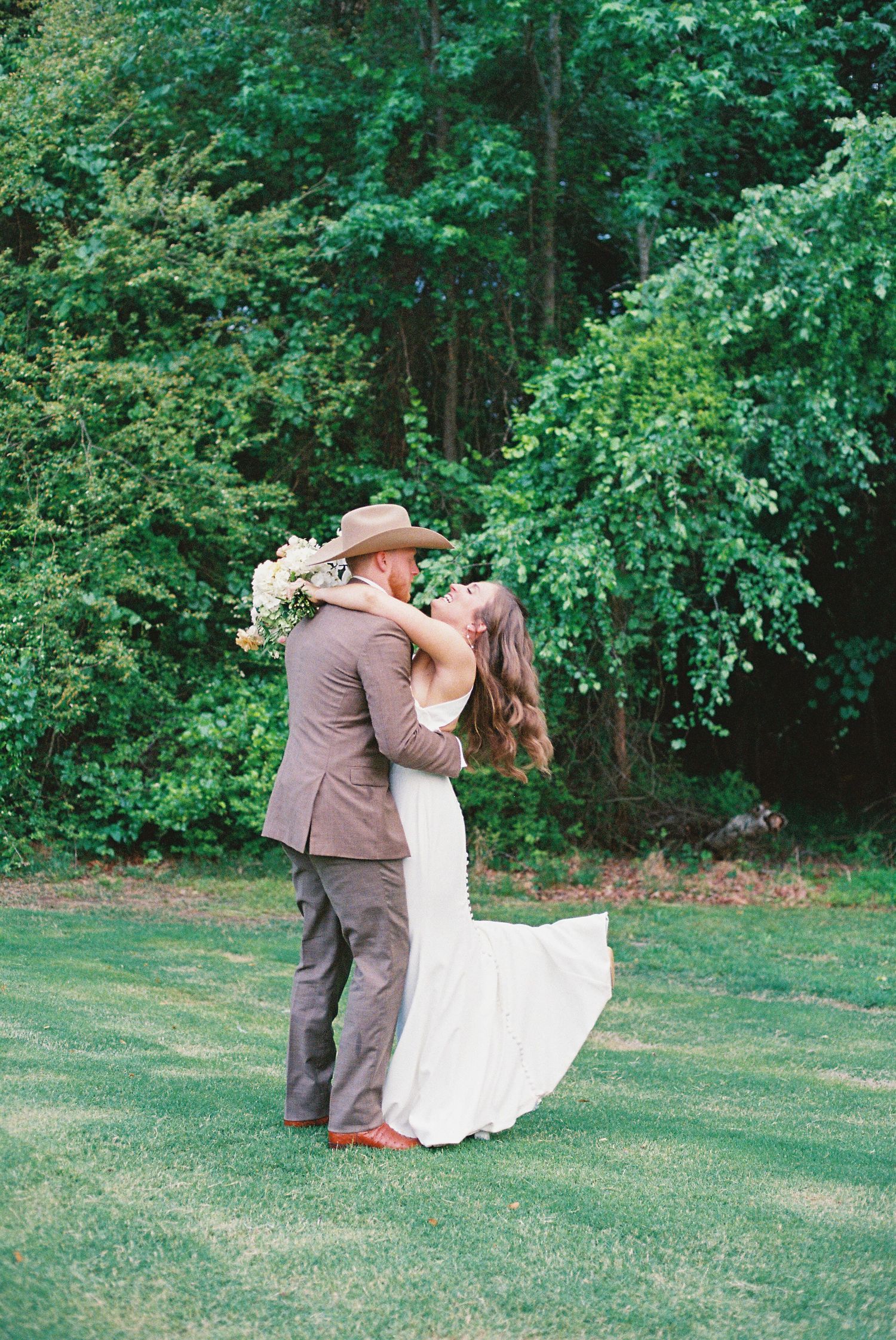 Bride and groom share a romantic kiss on green lawn with lush forest backdrop at outdoor wedding ceremony.