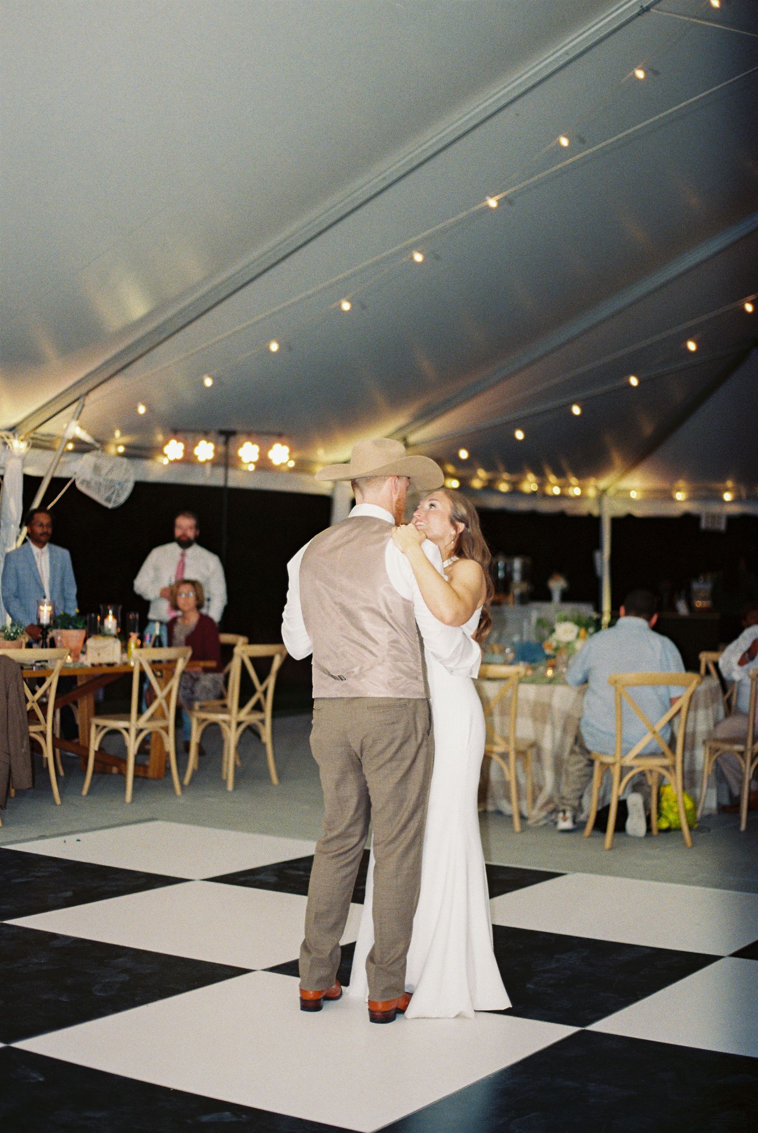 Bride and groom share first dance on checkered floor under a romantic tent with string lights at their wedding reception.