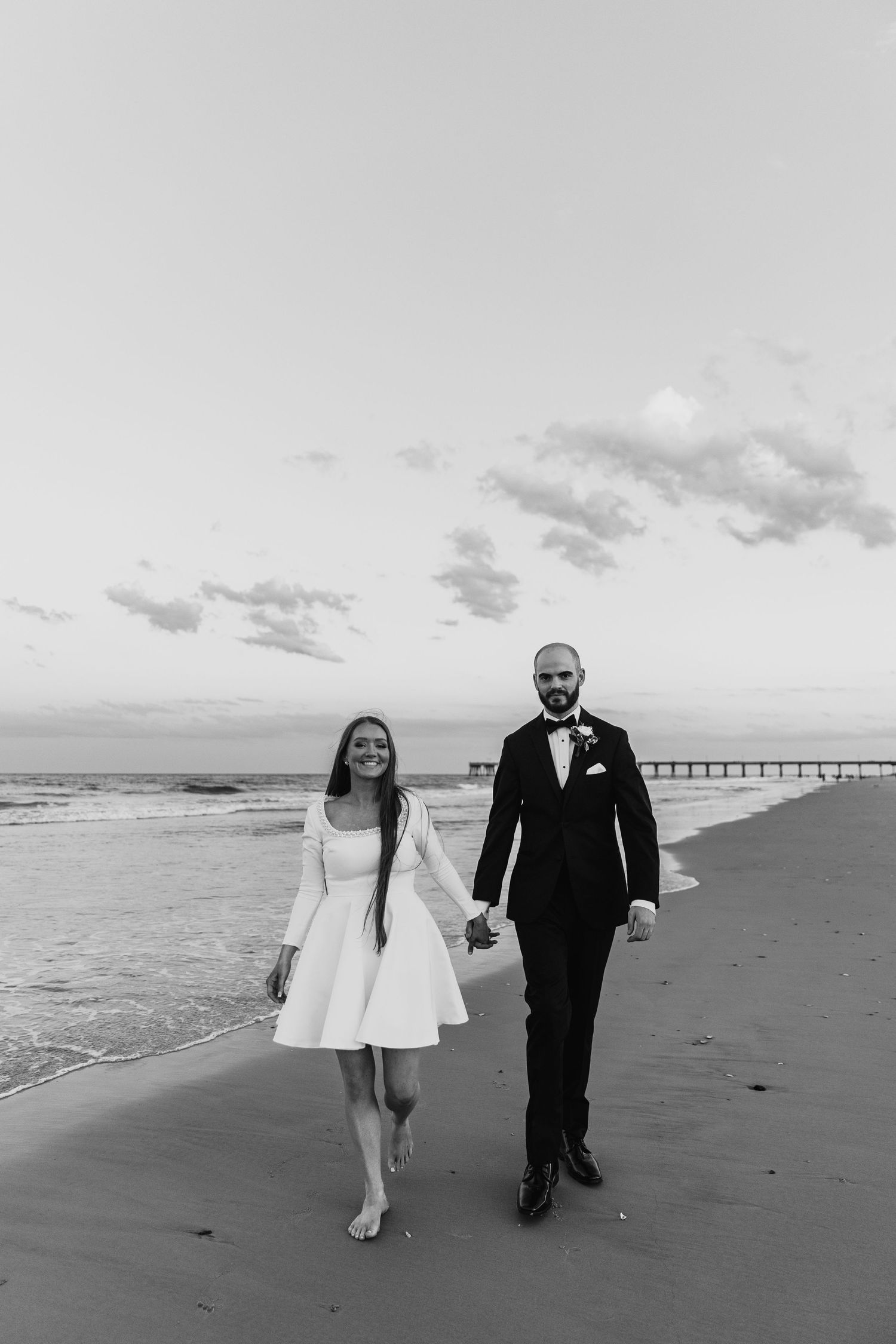 A black and white photo of a couple walking hand in hand on a beach with a pier in the background.
