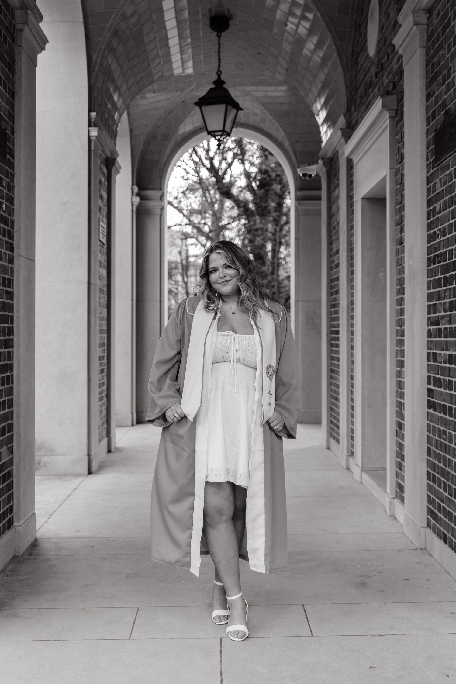 Black and white photo of graduate in cap and gown walking through elegant brick archway.
