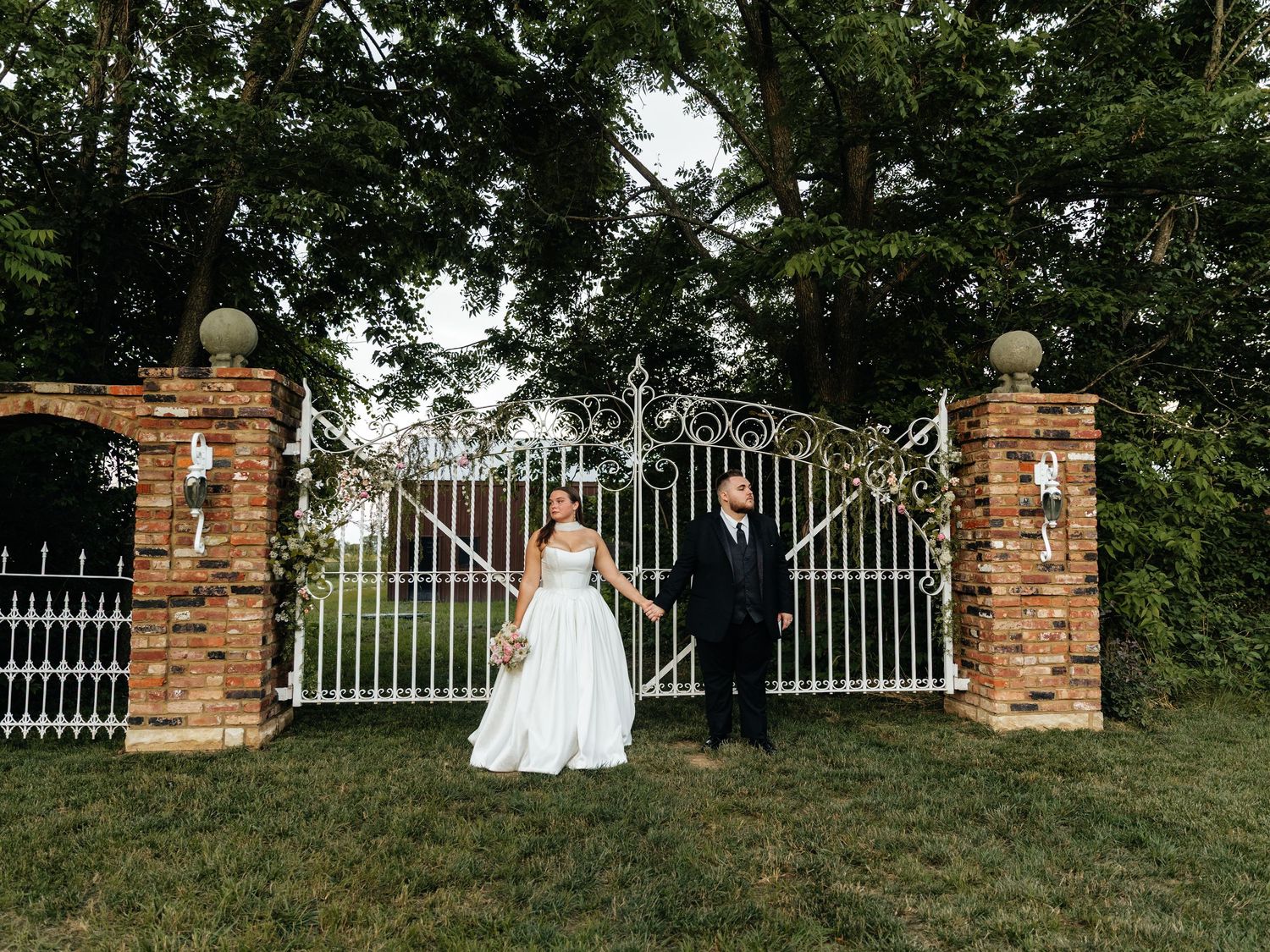 Wedding couple poses by decorative white iron gates and brick pillars on a grassy hillside.