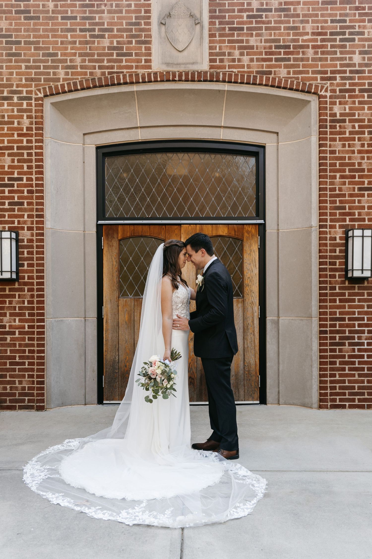 Wedding couple stands in elegant pose in front of brick archway entrance.