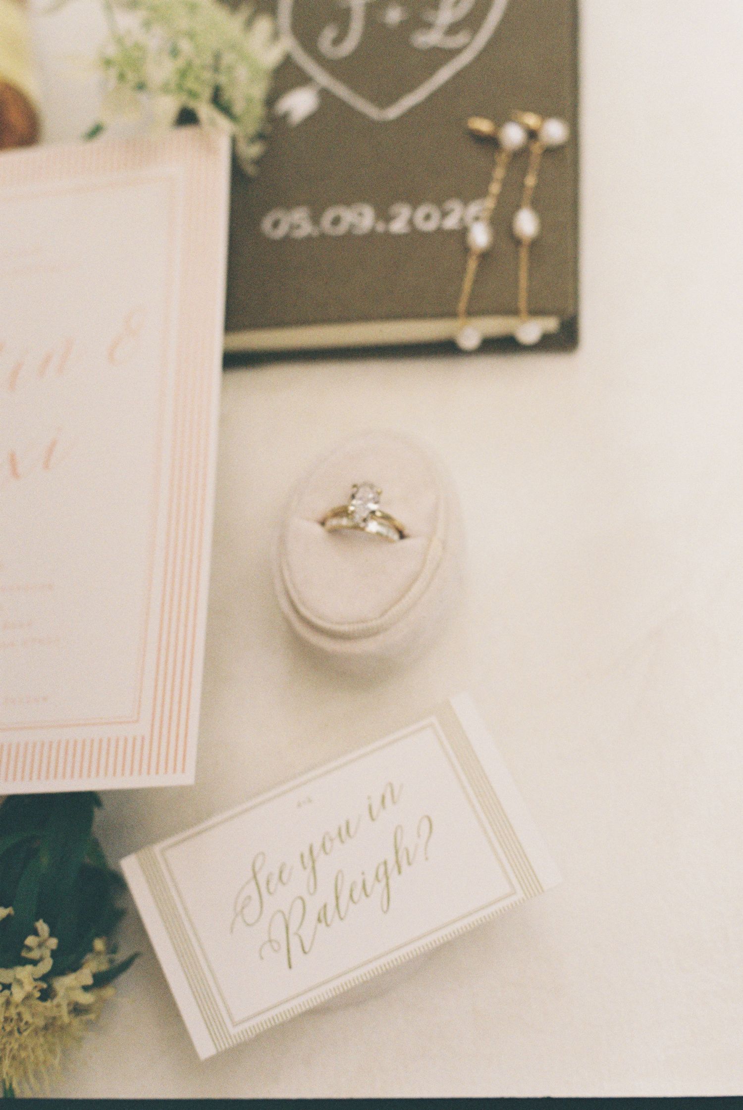 Elegant wedding flat lay with diamond engagement ring, save the date card, and Raleigh wedding invitation on white surface.