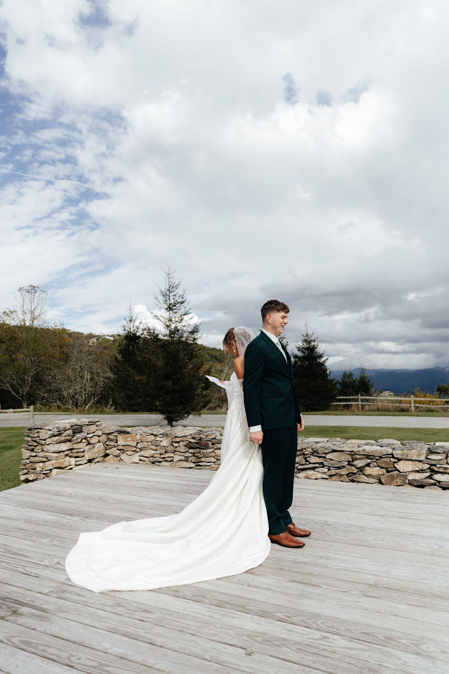 Bride in white gown with long train stands back-to-back with groom in dark suit on wooden deck at outdoor wedding venue.