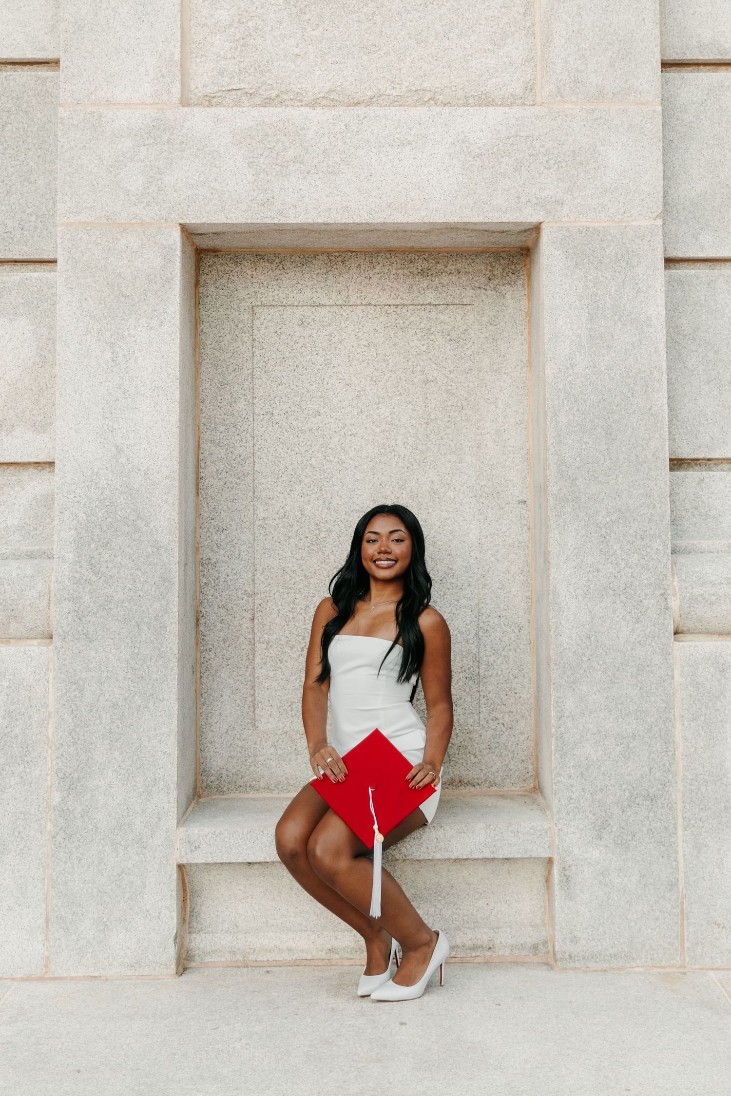 Smiling young woman in white dress holding red graduation cap posed in stone wall niche.