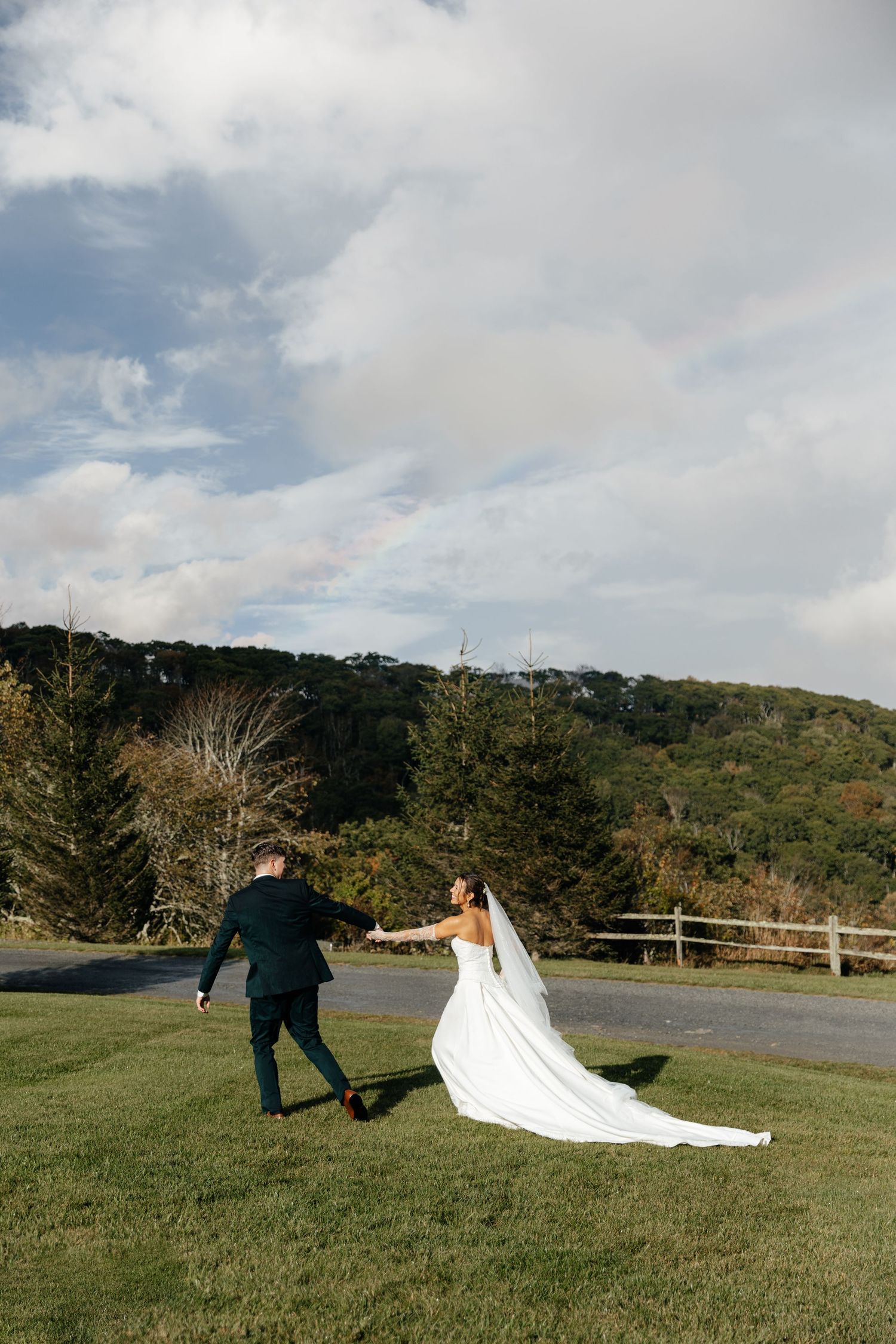 Bride and groom walking hand in hand across green lawn with rolling hills and cloudy sky backdrop.