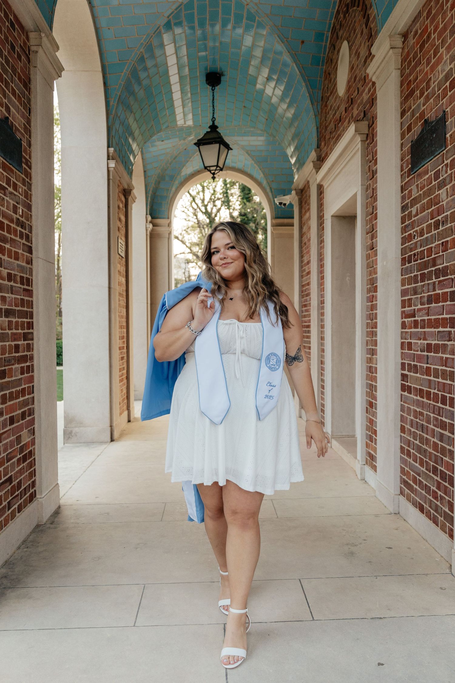 A smiling graduate in a white dress holds a blue gown over her shoulder in a brick archway corridor.