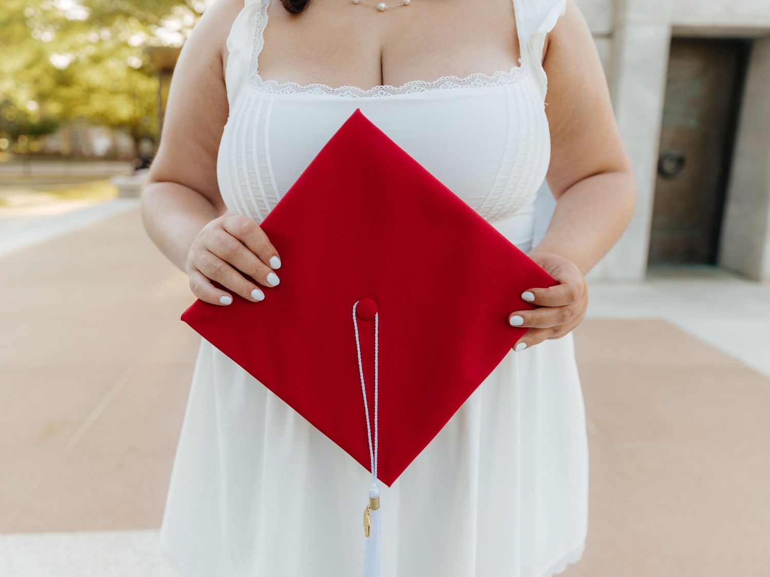 Woman in white dress holding a red graduation cap, celebrating academic achievement outdoors.