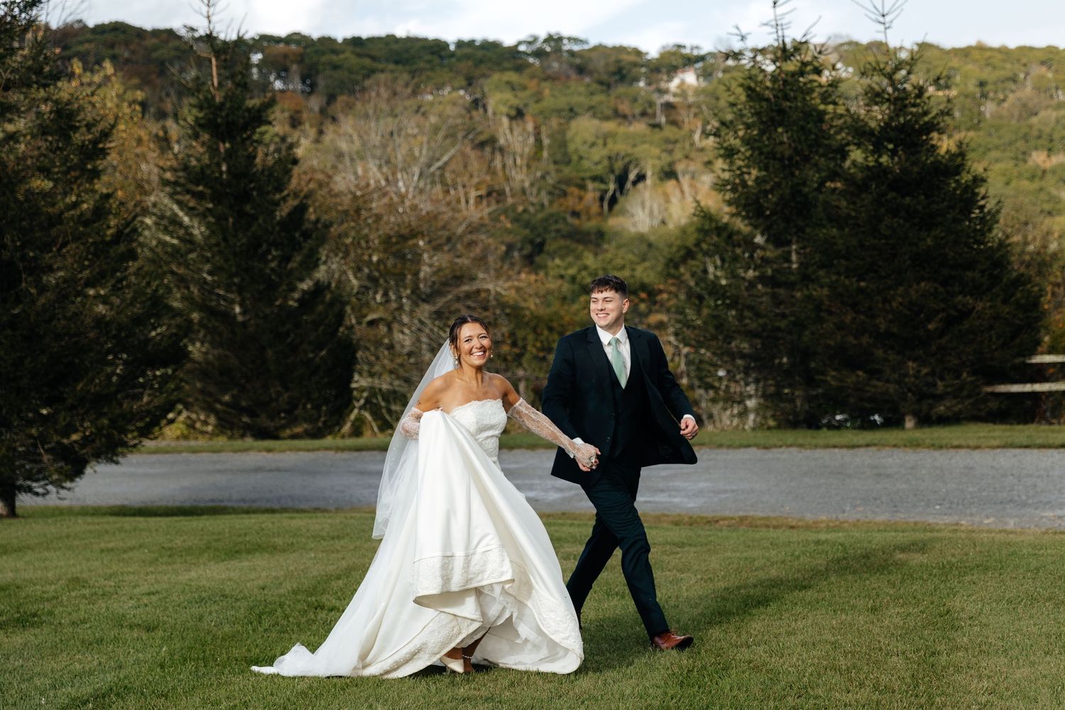 Bride and groom holding hands walking on green lawn with trees in background on their wedding day.