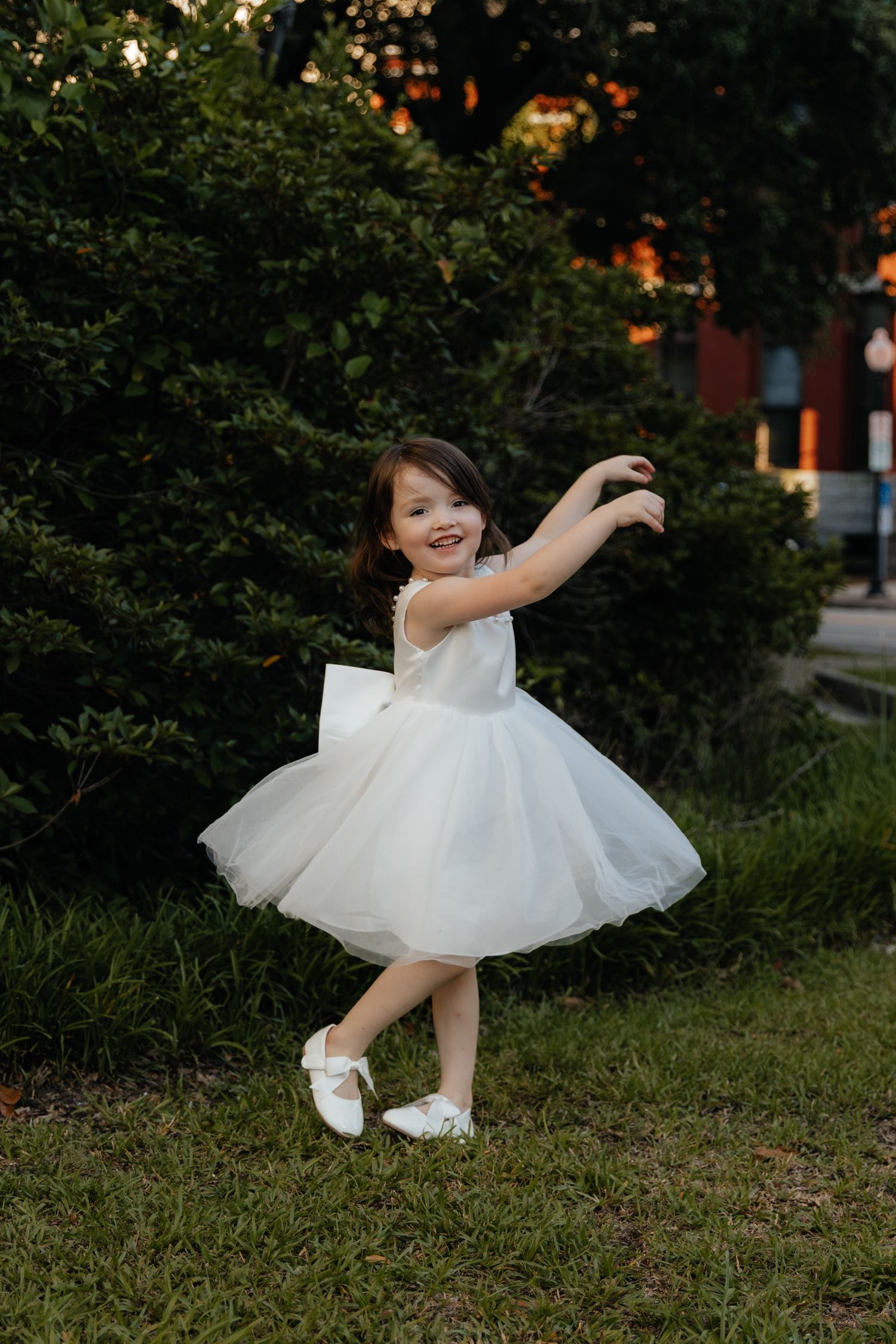 A young flower girl in a white tulle dress twirls joyfully in a garden setting.