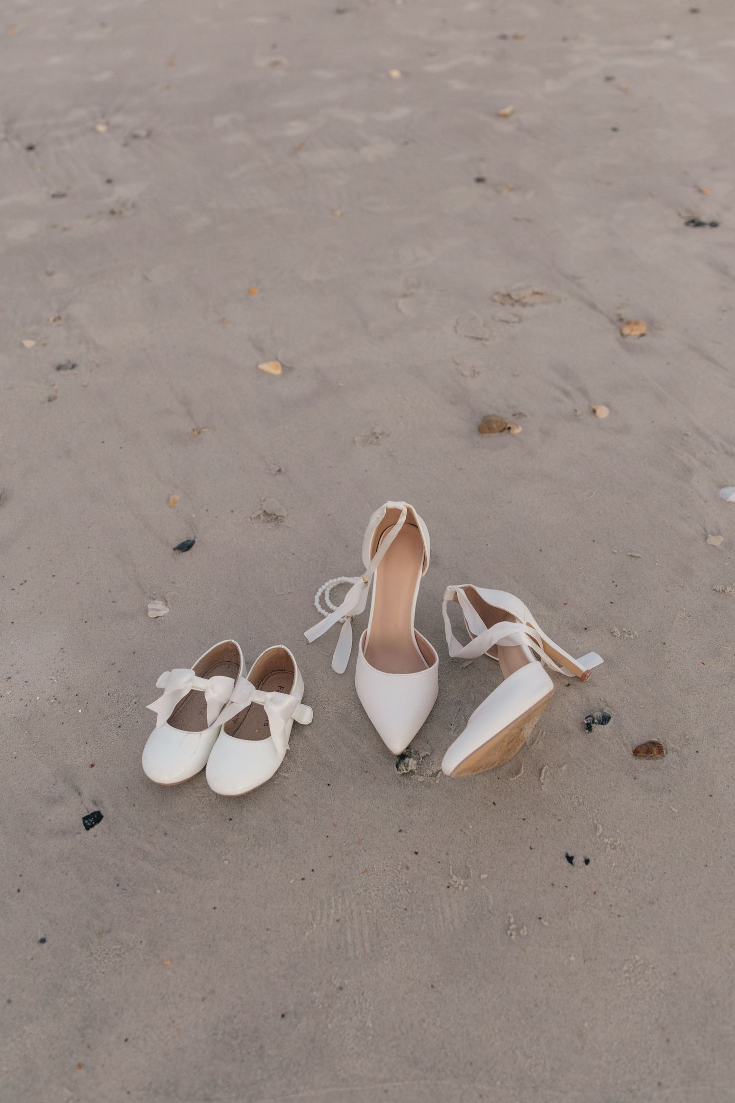 Wedding detail shot of two pairs of white shoes arranged on sandy beach.