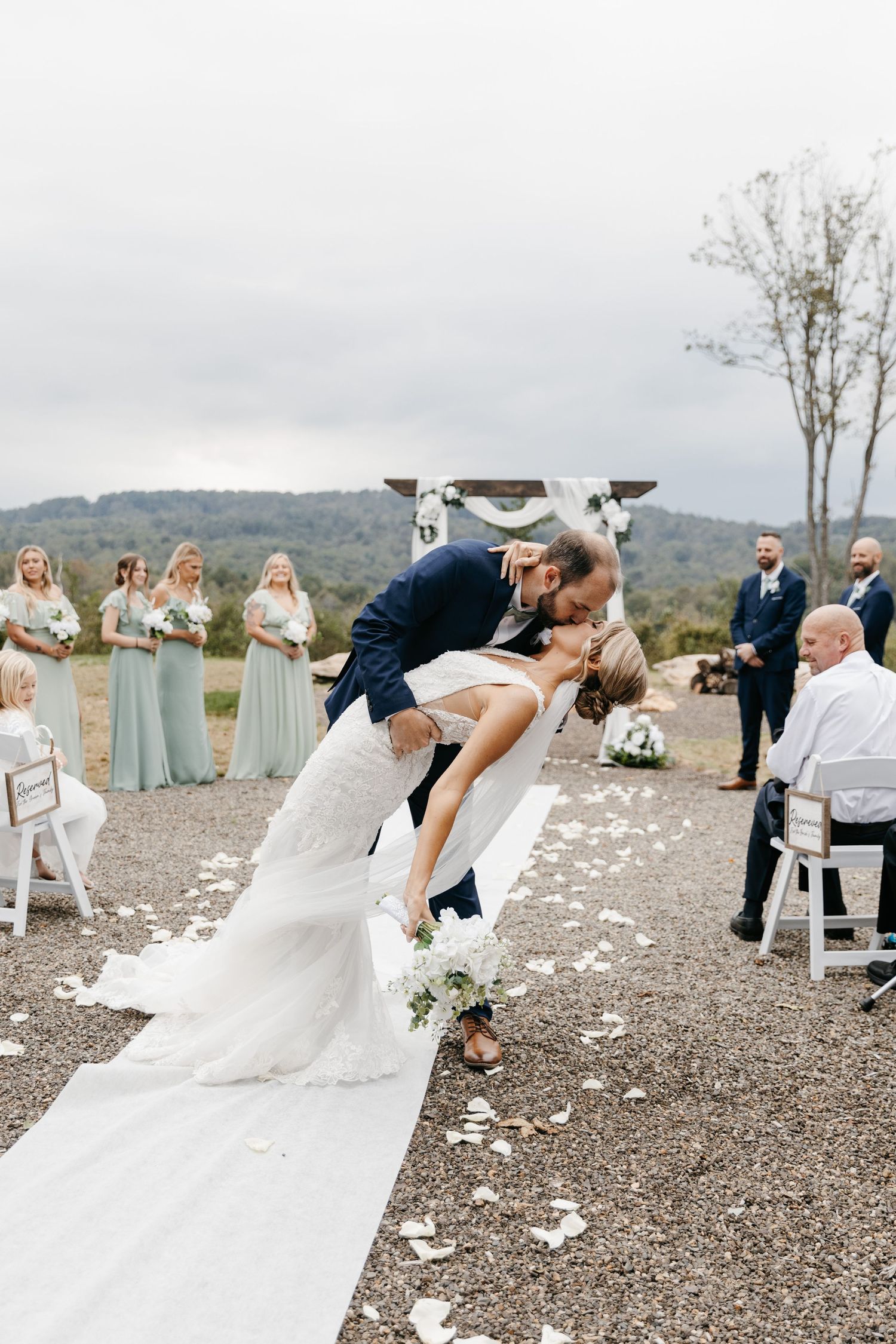 Groom dips bride for a romantic kiss at outdoor wedding ceremony with bridesmaids in sage green dresses.