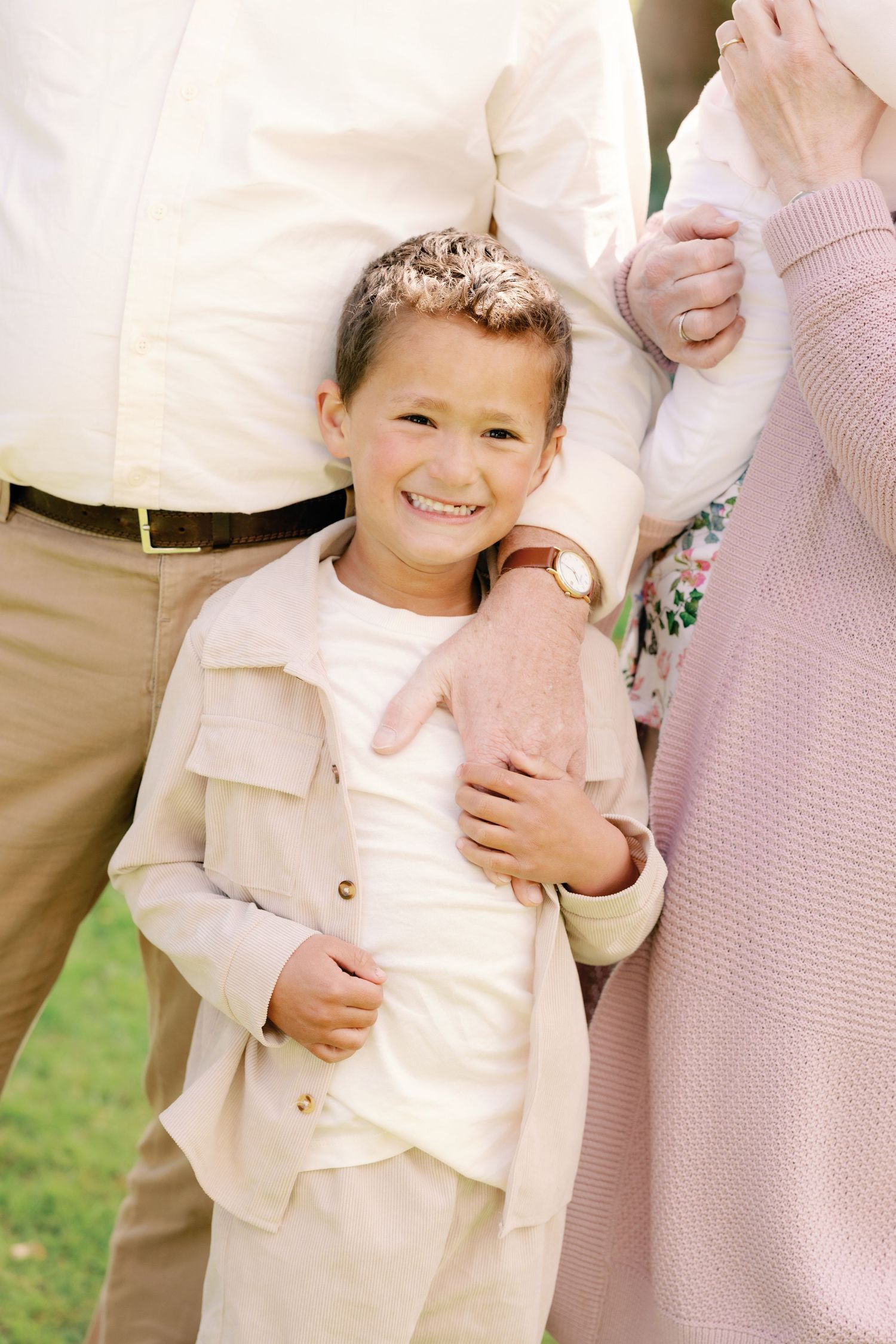 Smiling young boy in beige outfit standing between parents outdoors, being embraced warmly on a sunny day.