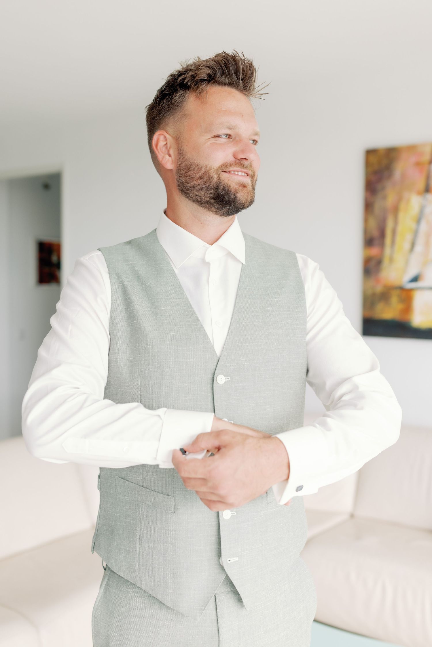 A person wearing a light gray vest and white dress shirt adjusts their cuff in an elegant indoor setting.