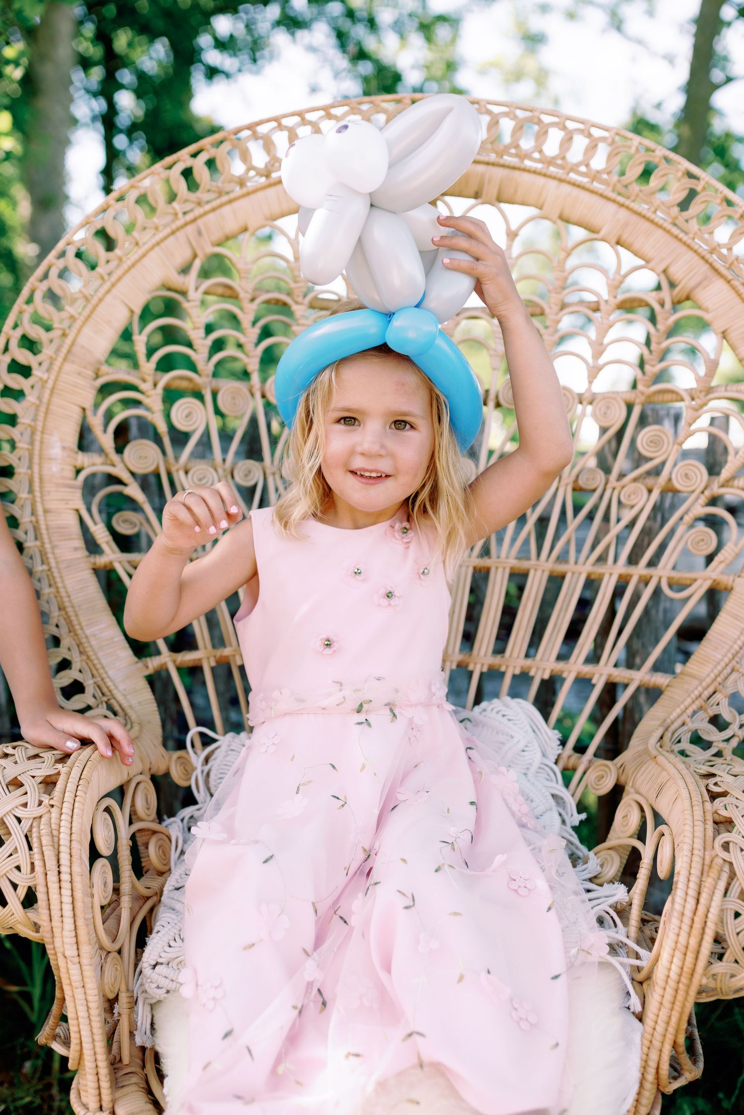 A child in a pink dress poses playfully on a wicker peacock chair in an outdoor garden setting.
