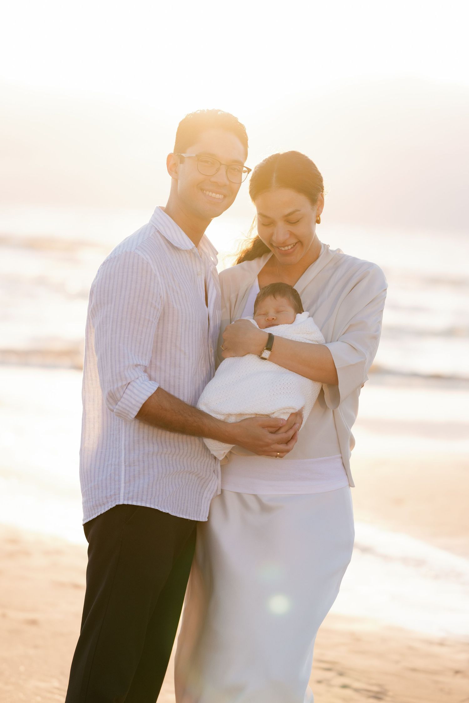 A couple stands on a beach at sunset holding a newborn baby wrapped in white, with soft golden light illuminating the scene.