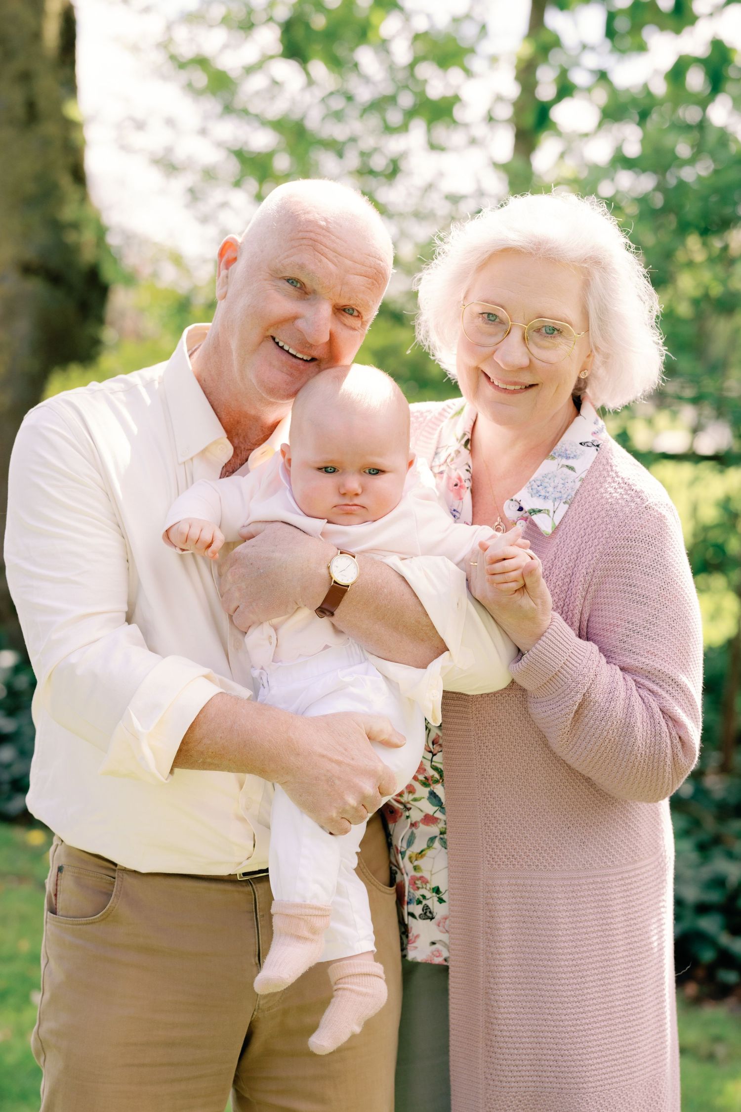 Smiling grandparents holding a cute baby outdoors in a sunny green garden setting.