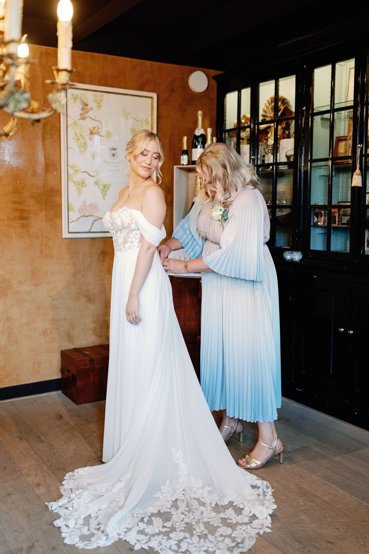 Two women in elegant dresses prepare for a wedding, with the bride wearing a flowing white gown with lace details.