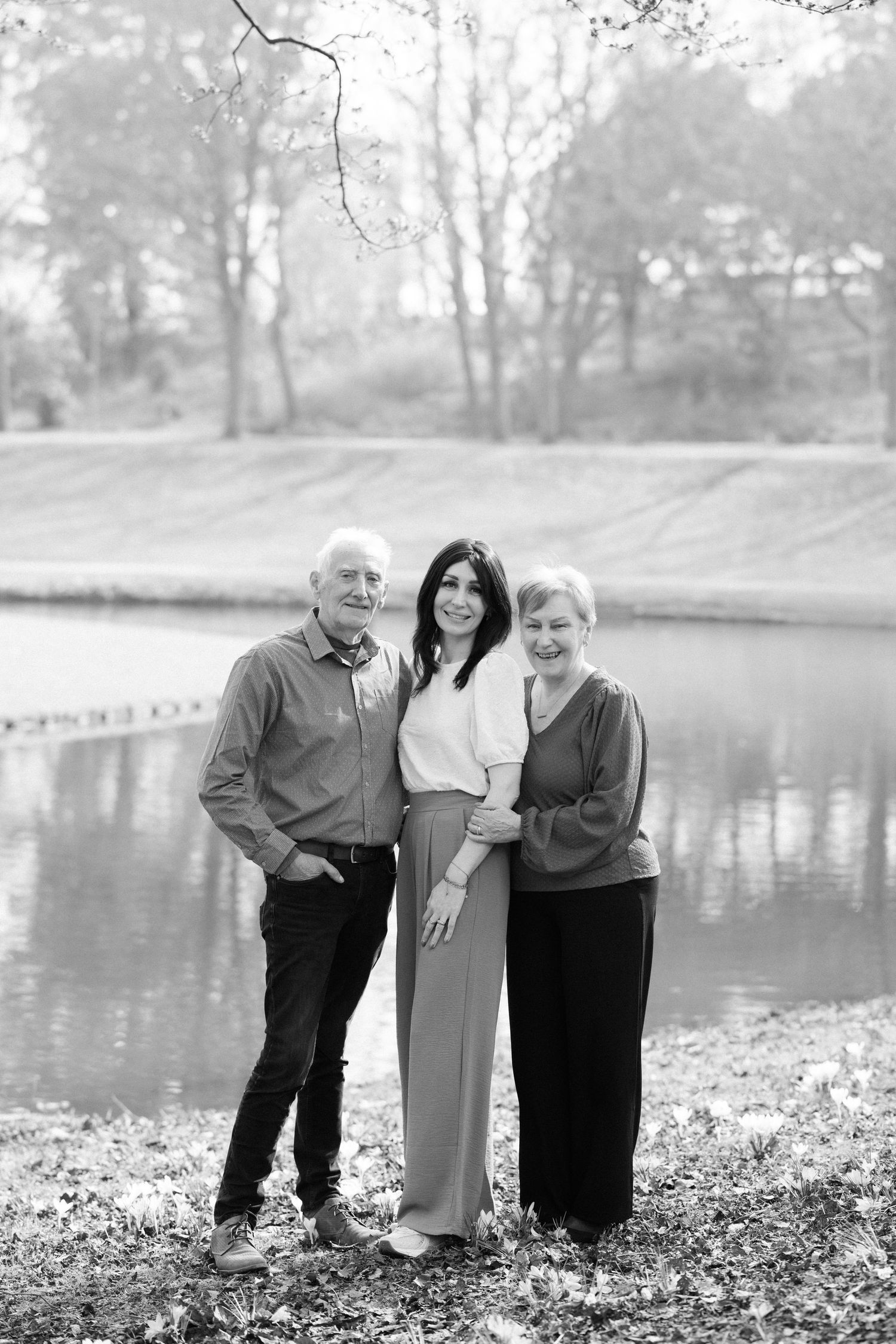 Three people posing together by a lake on an autumn day with fallen leaves on the ground.
